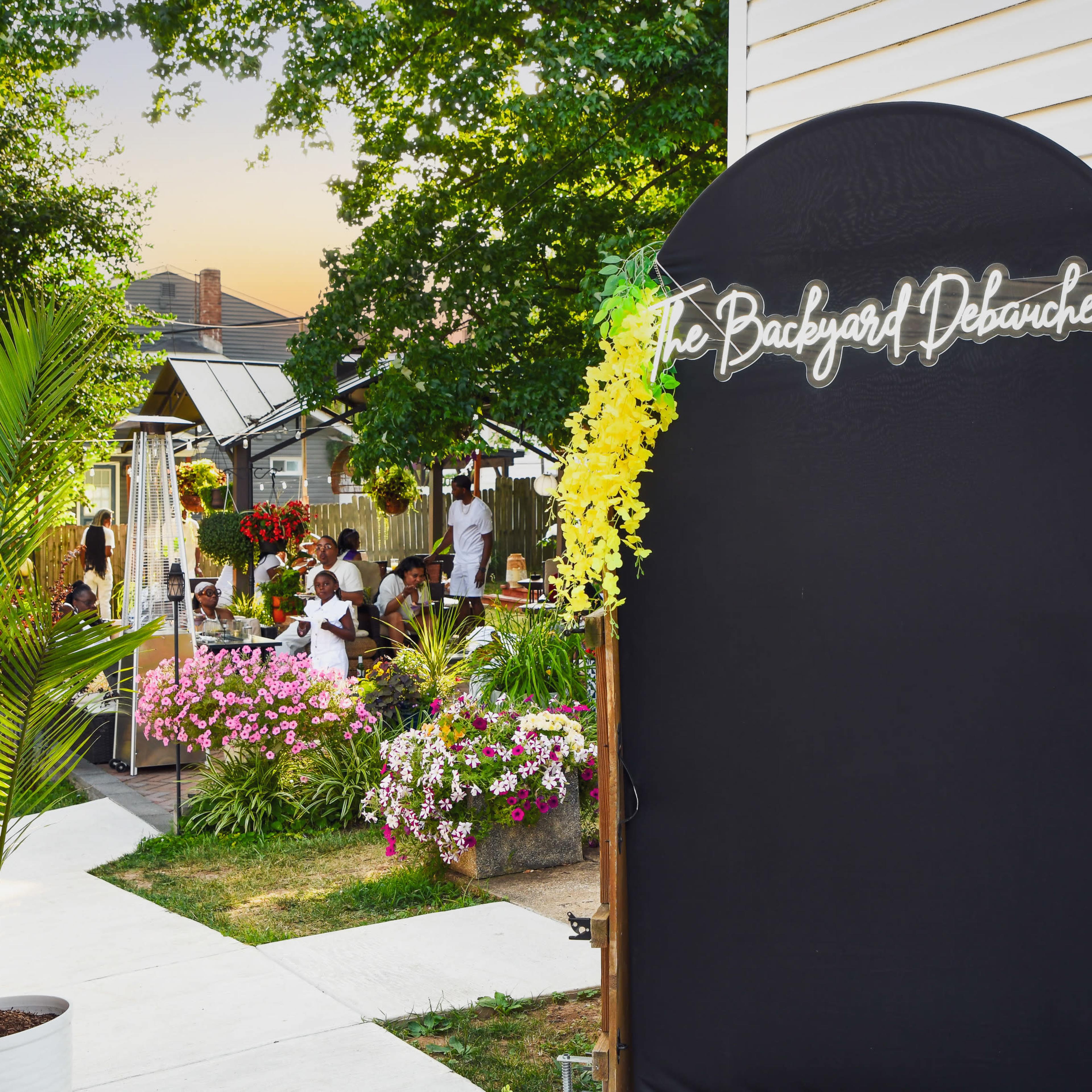The scene shows an outdoor gathering area with a sign reading "The Backyard Debauchery," surrounded by colorful flowers and greenery.