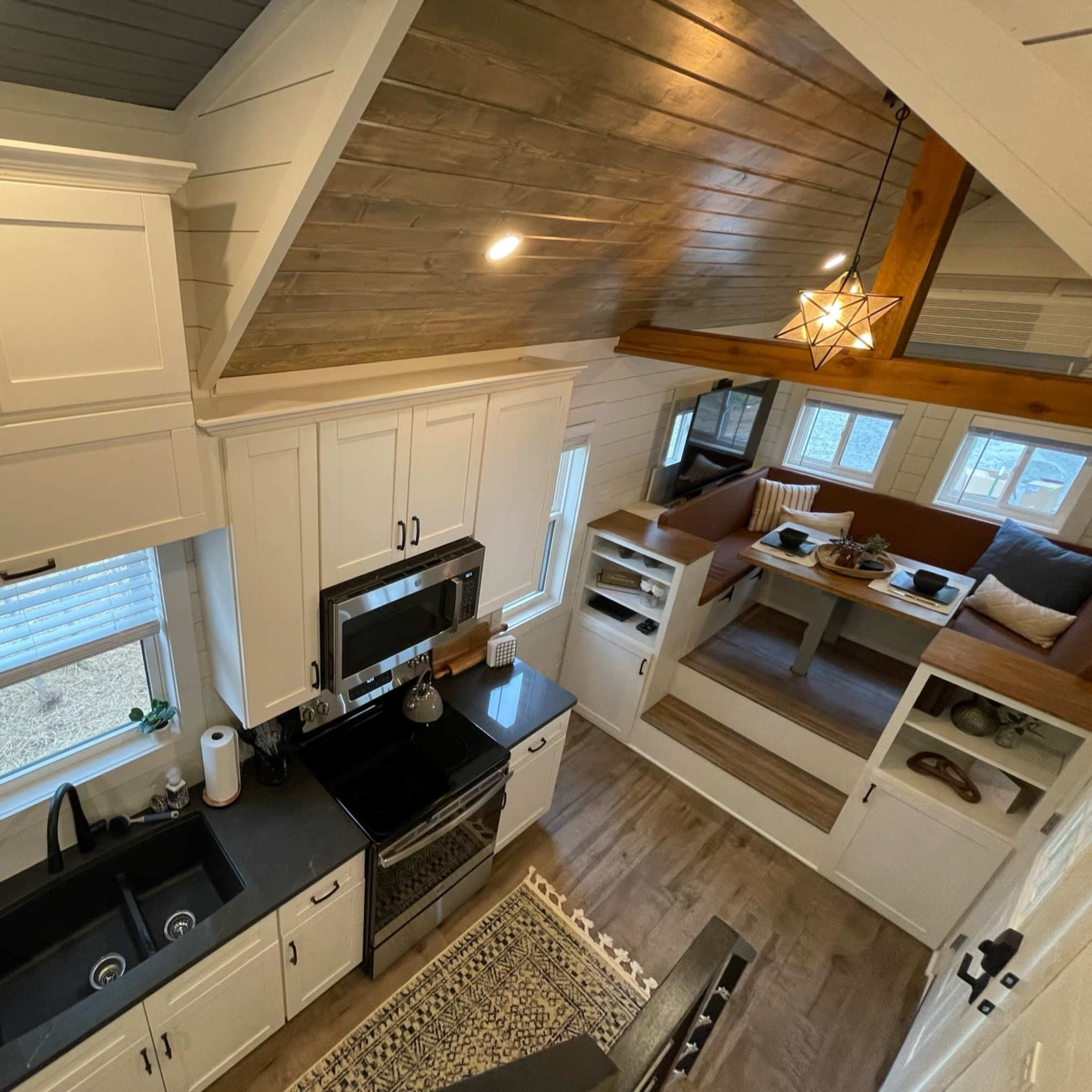 A small, modern kitchen with white cabinetry, black countertops, and a cozy seating area featuring built-in shelves and large windows.