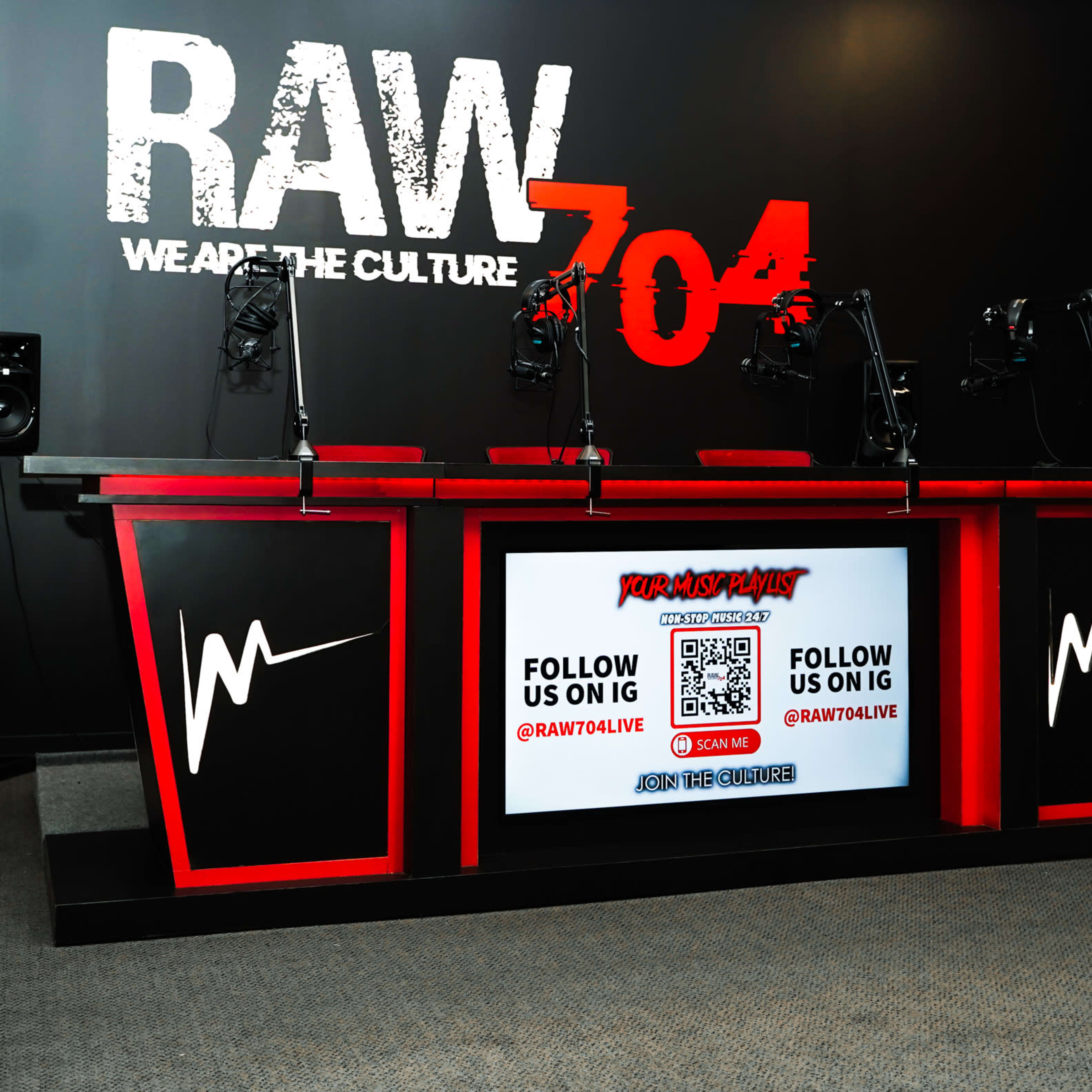 A modern studio setup with a red and black broadcast table featuring multiple microphones and a large screen displaying social media information against a black wall with bold text.