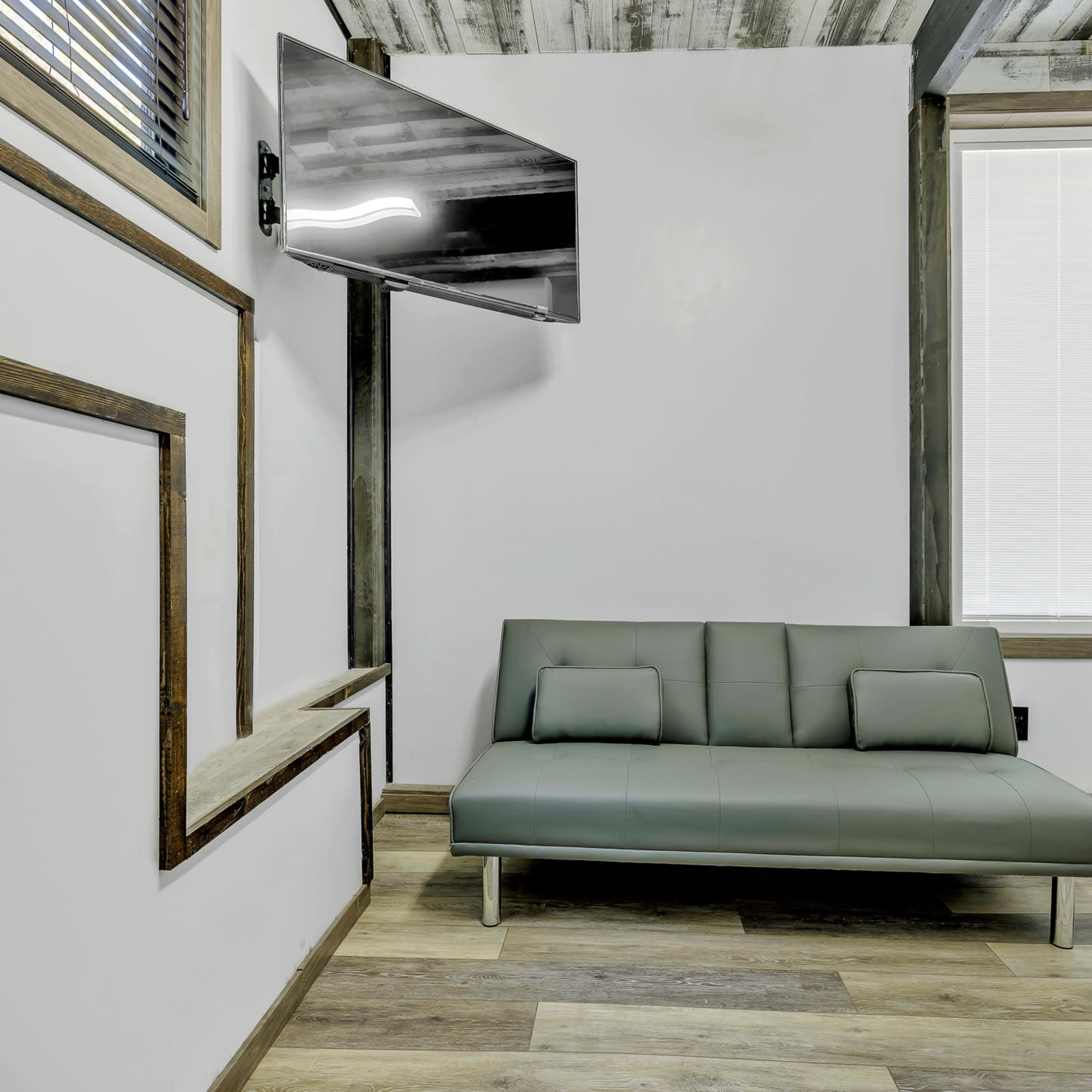 A minimalist room features a green sofa bed, a wall-mounted television, and a large window with blinds.