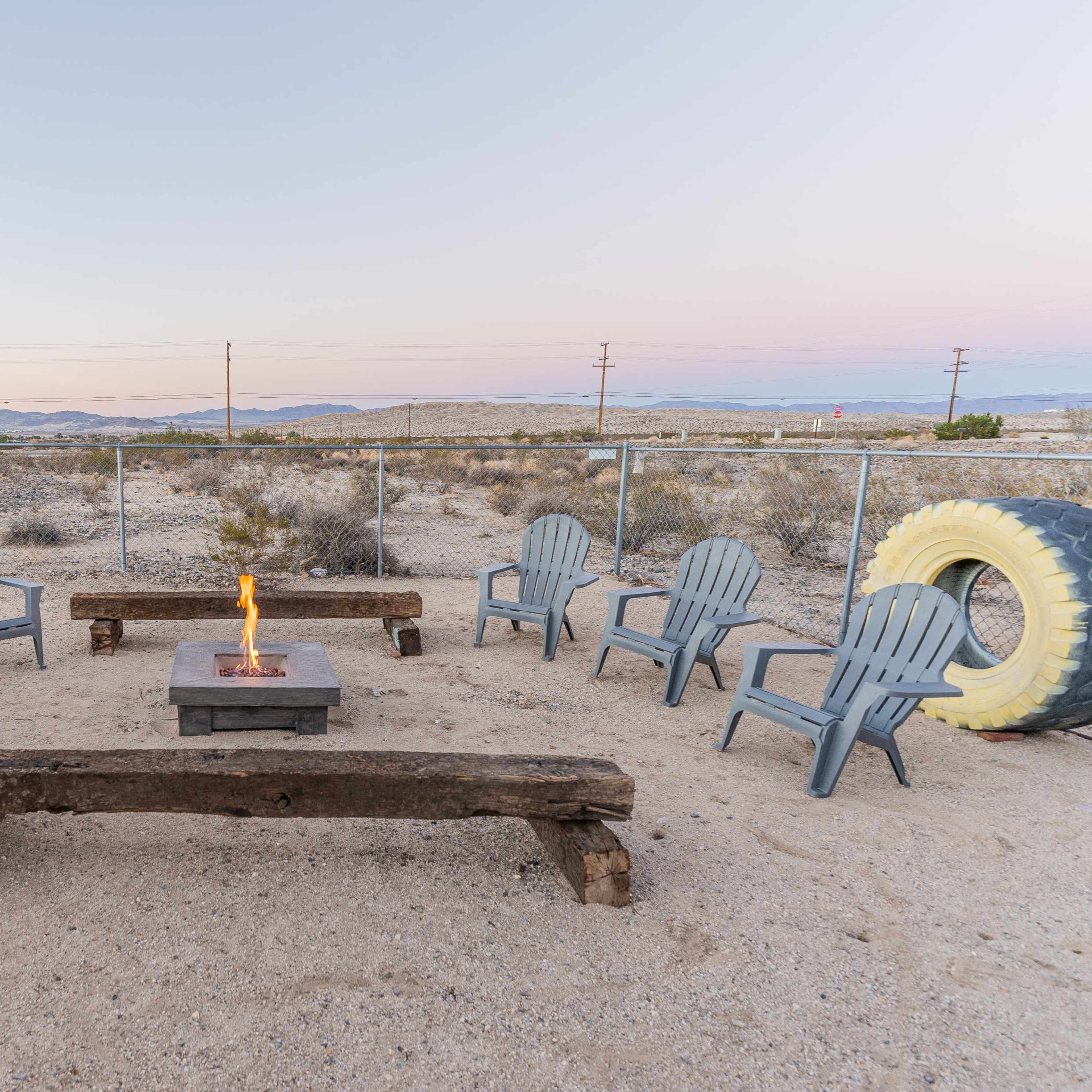 A circular seating area made of gray chairs surrounds a fire pit with a wooden bench, set in a desert landscape with a large tire visible nearby.