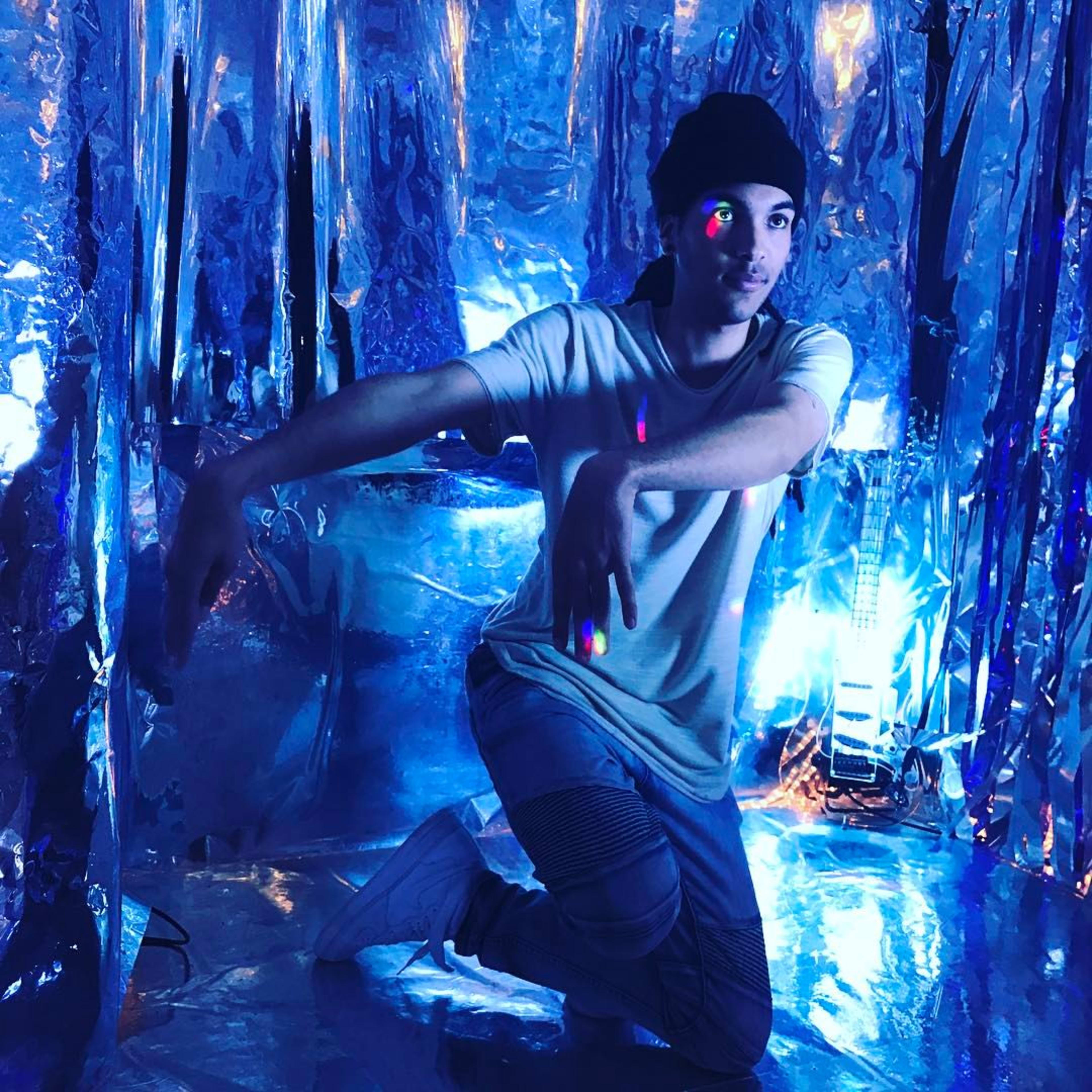 A person wearing a light-colored shirt and a beanie poses in a reflective, blue-toned environment with colorful light reflections.