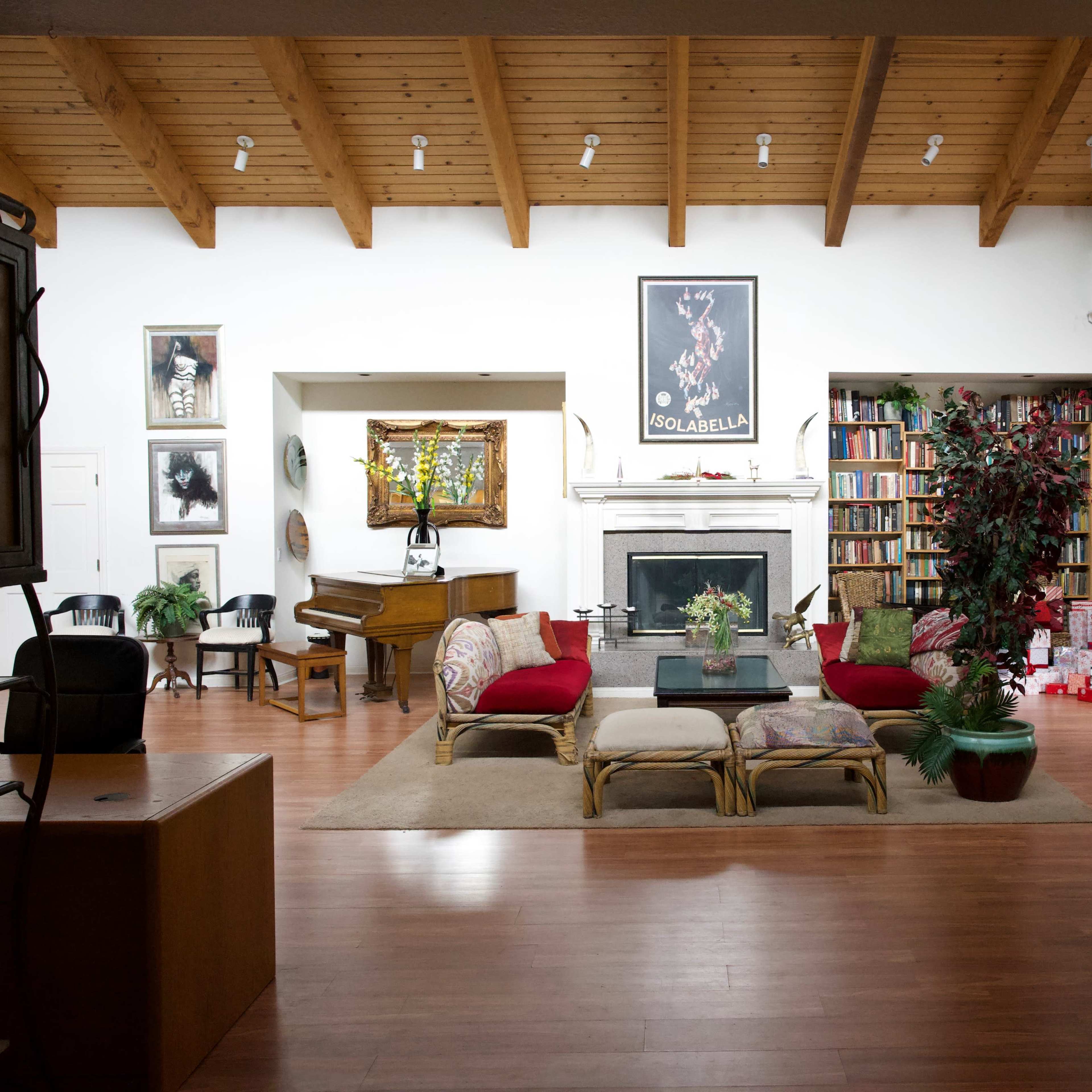 A spacious living room with a high wooden ceiling, a large fireplace, bookshelves filled with books, a Christmas tree, and a grand piano.