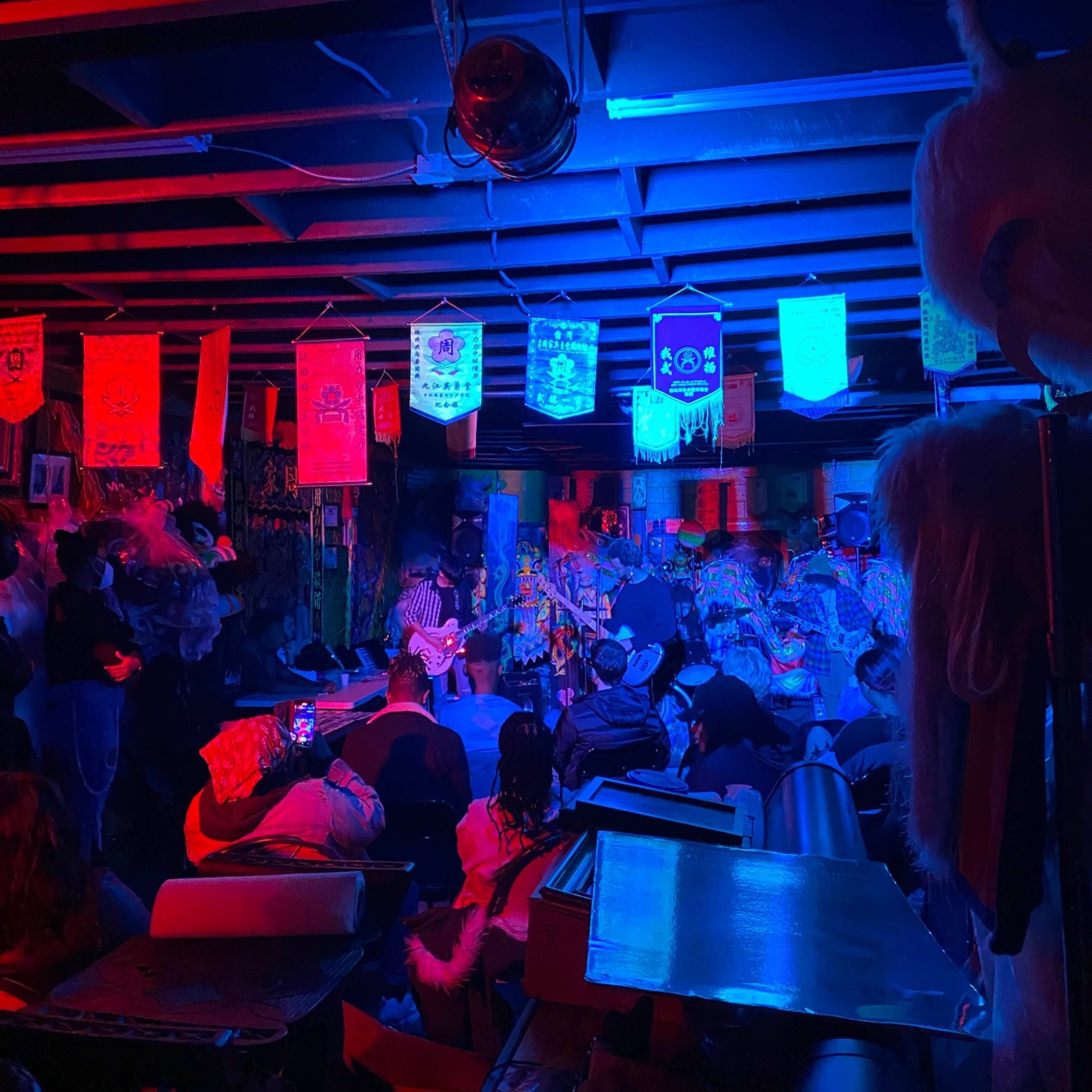 A dimly lit basement venue filled with an audience seated and facing performers surrounded by colorful banners and vibrant decor.