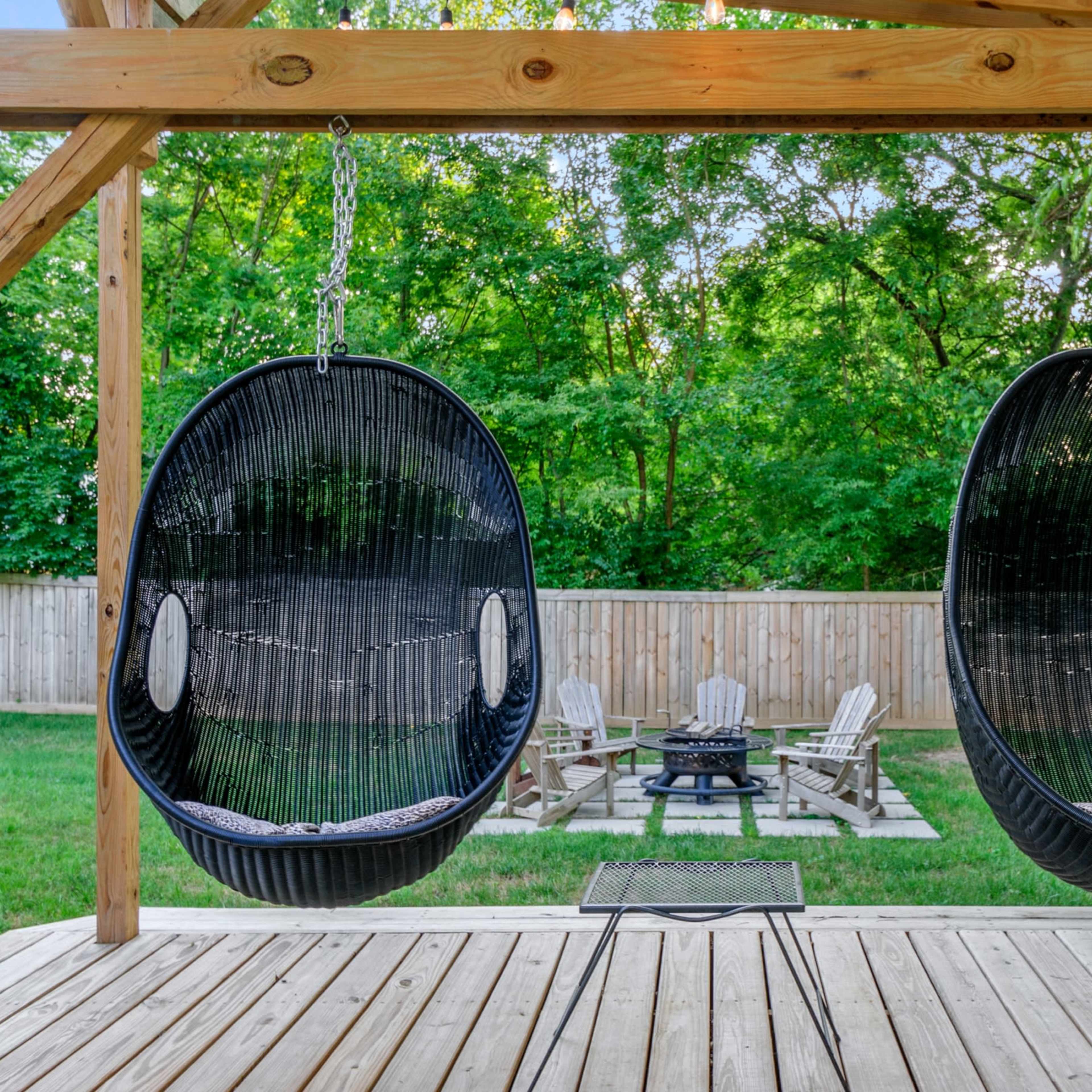 Two hanging chairs on a wooden deck overlooking a grassy area with a fire pit and Adirondack chairs in the background.