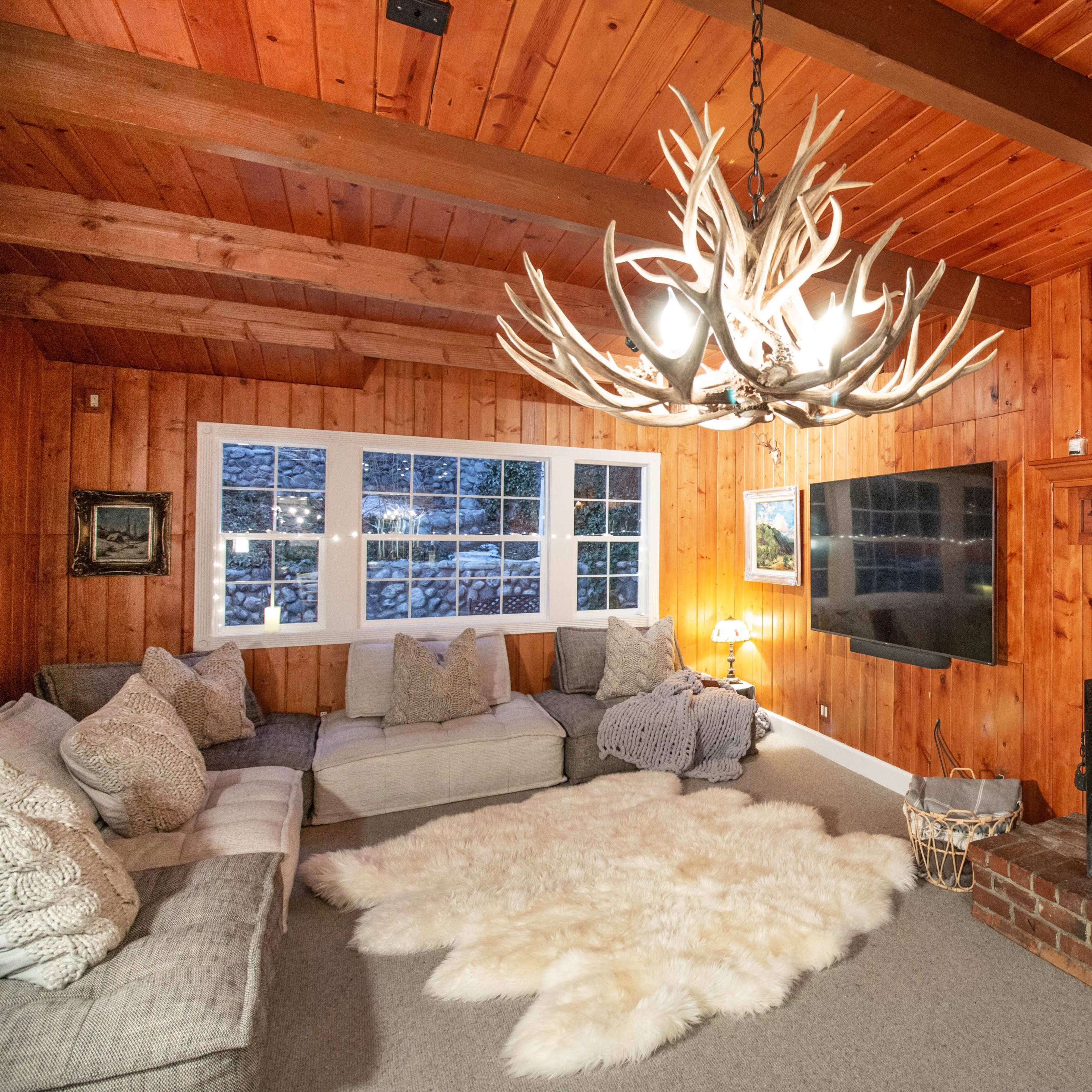 A cozy living room with wooden walls, a brick fireplace, a large antler chandelier, and a plush area rug in front of a sectional sofa.