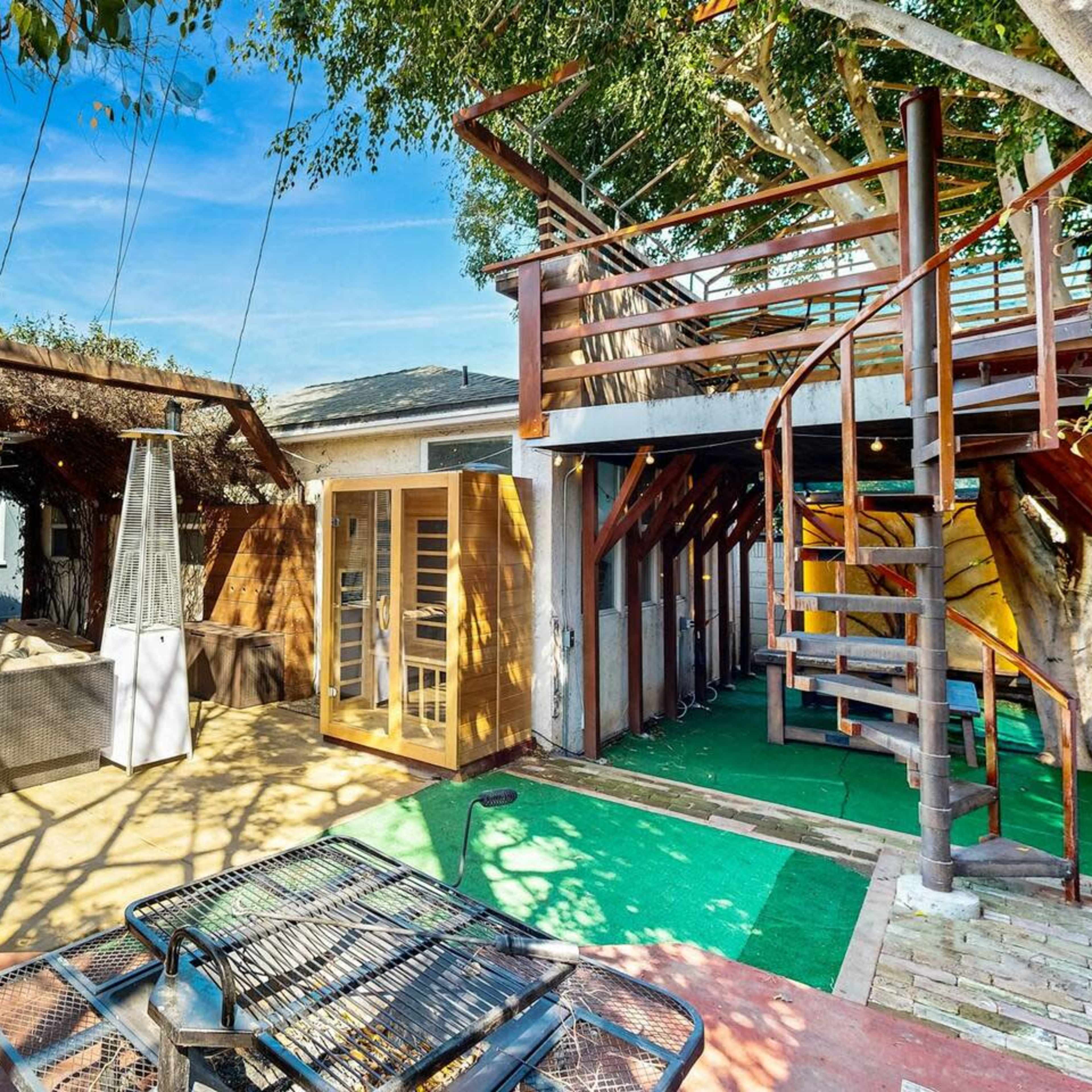 A backyard area featuring a covered patio with seating, a spiral staircase leading to an upper deck, and a wooden sauna alongside potted plants.