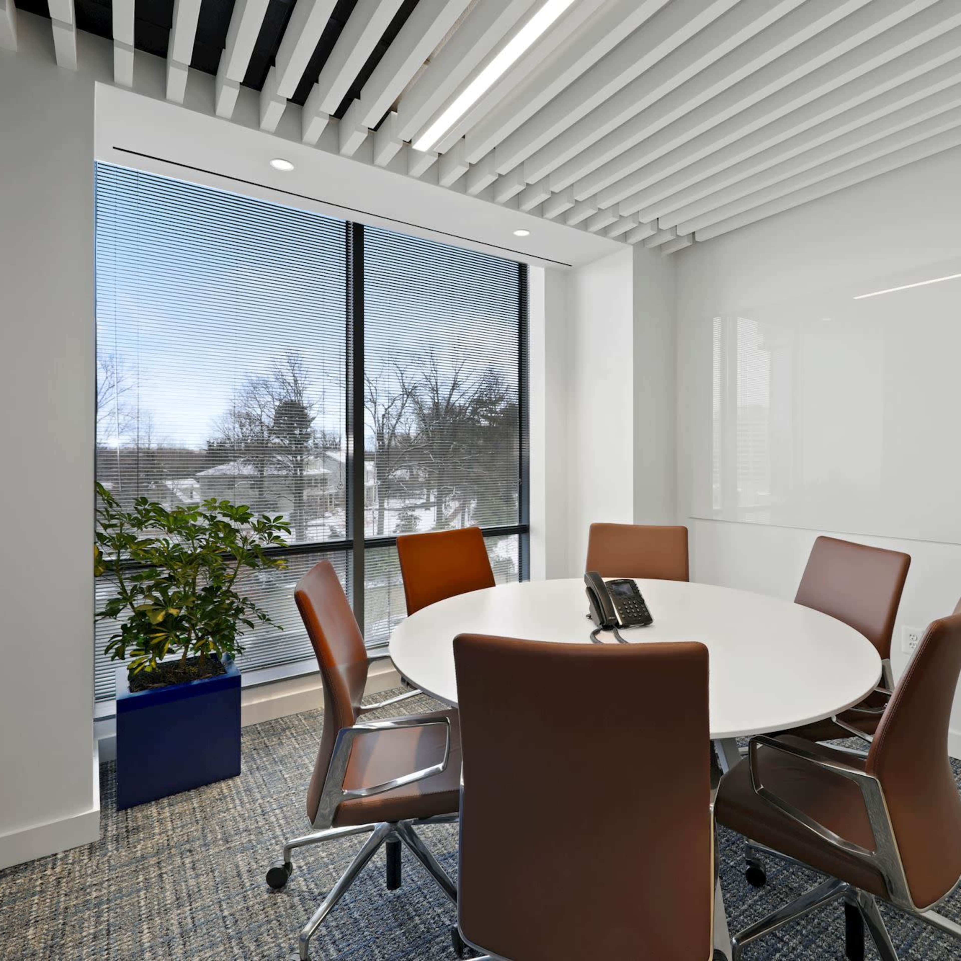 A modern conference room features a circular table surrounded by brown chairs, large windows with blinds, and a potted plant in a blue planter.