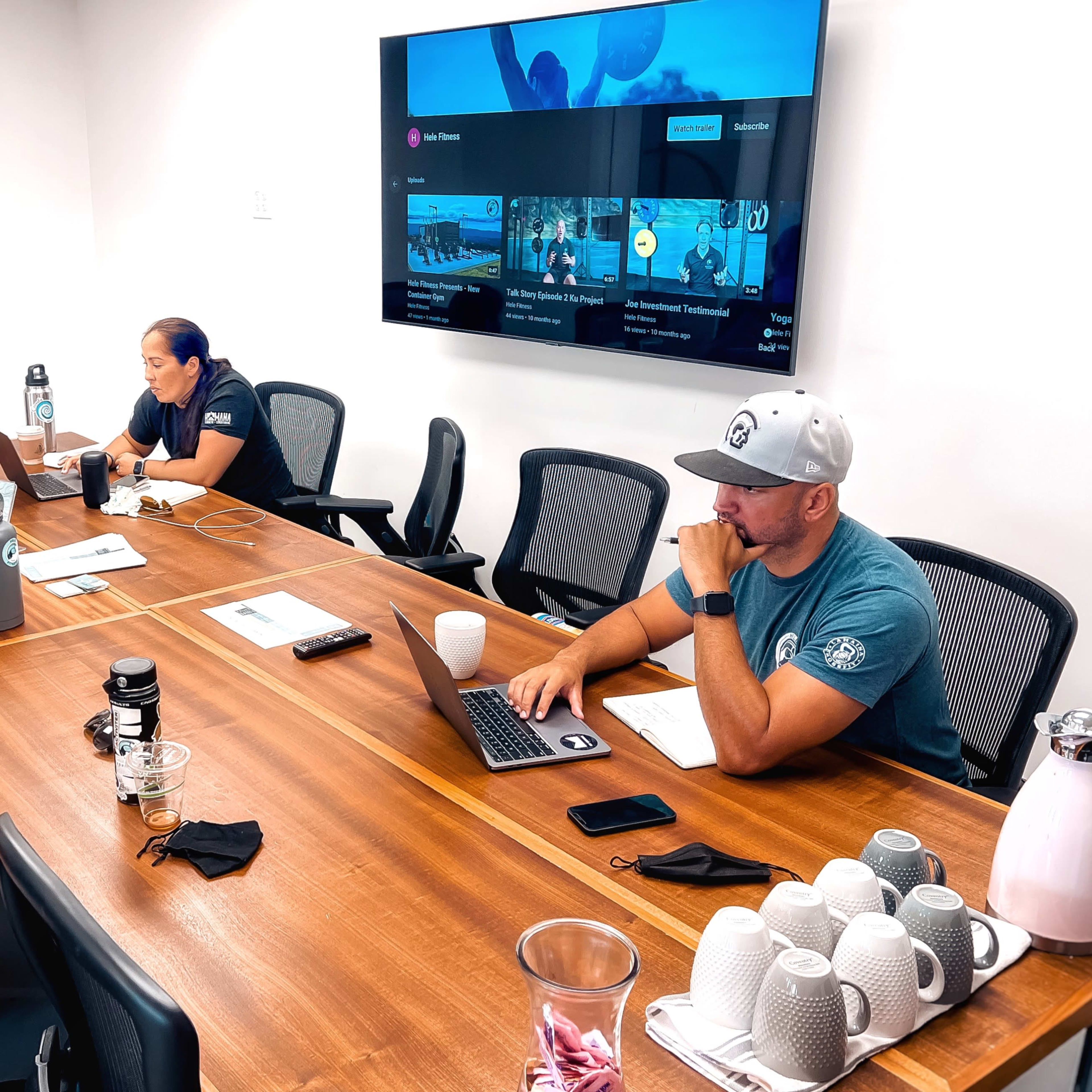 Four people are seated around a large wooden conference table in a meeting room, with a screen displaying content mounted on the wall.