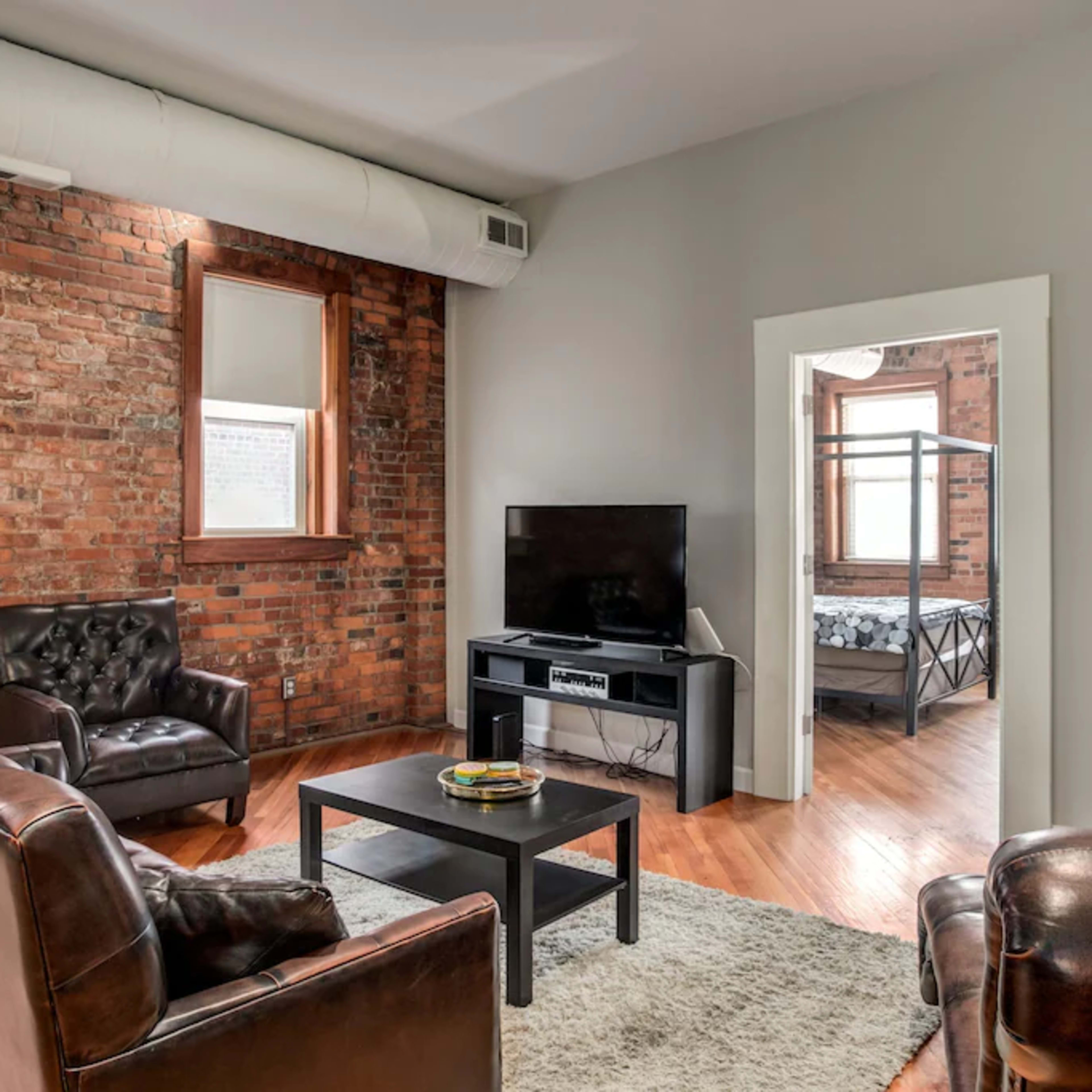 A cozy living room with exposed brick walls, two leather armchairs, a black coffee table, and a television against a wall leading into a bedroom visible through an open door.