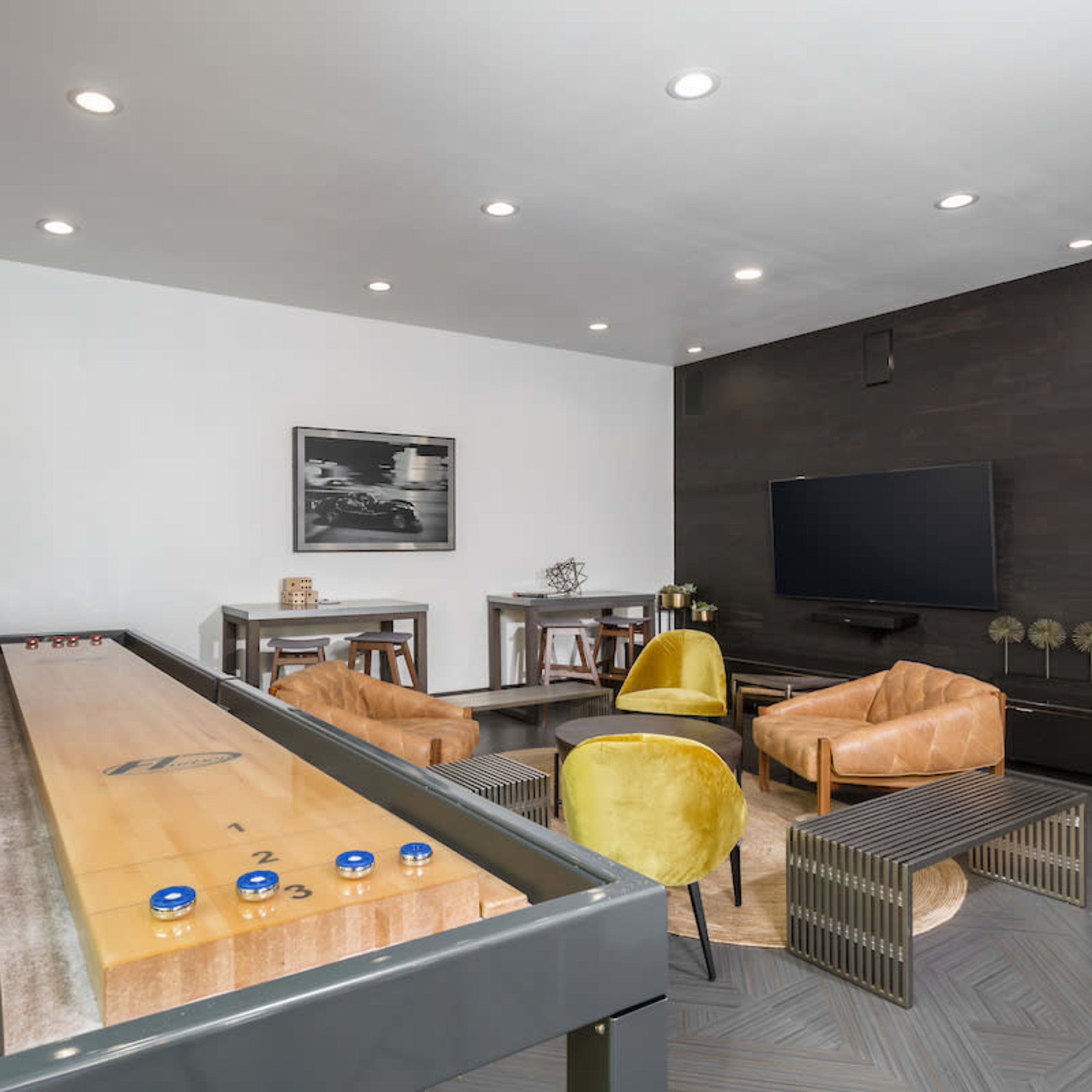 A modern game room featuring a shuffleboard table, two yellow chairs, and a wall-mounted television.