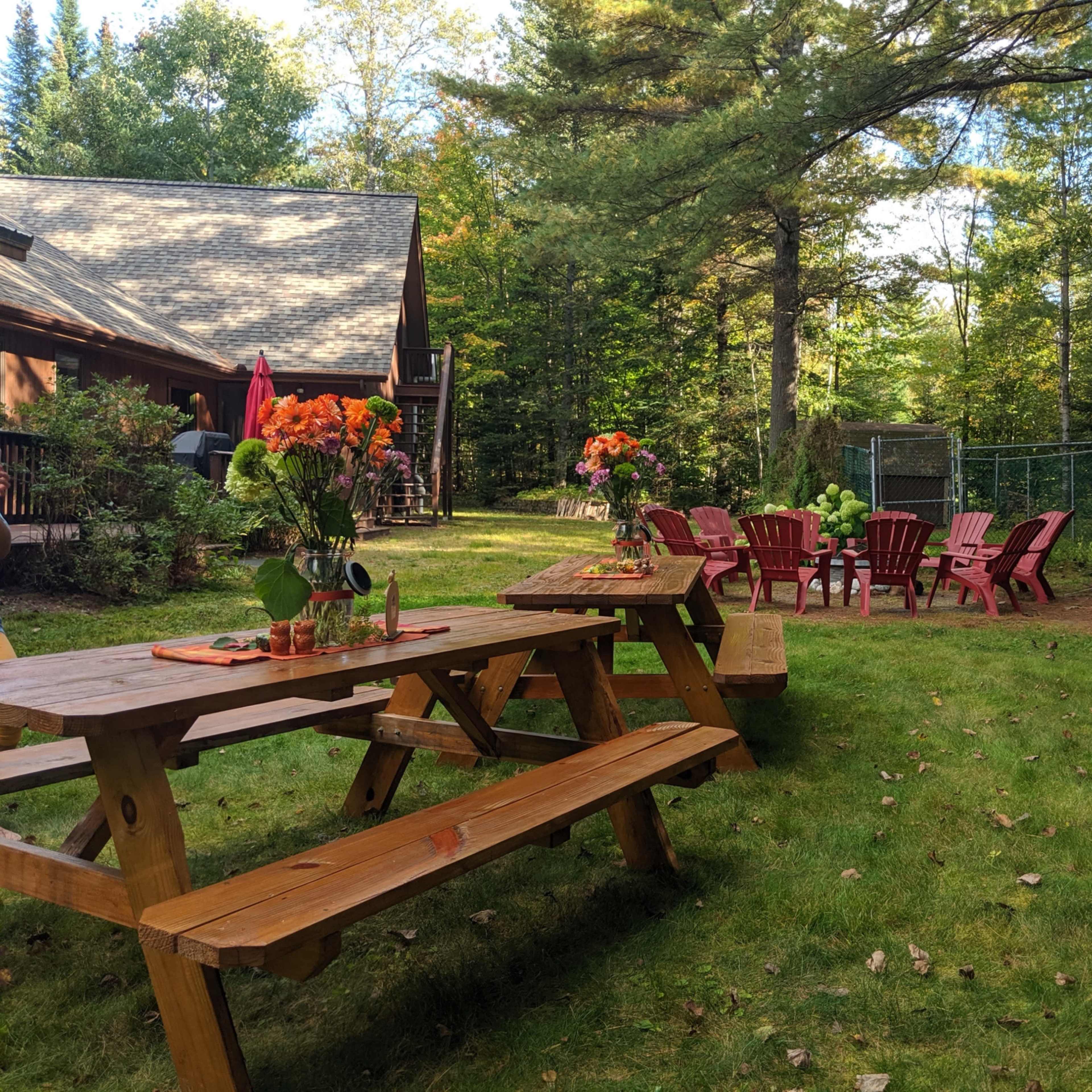 A backyard scene features wooden picnic tables and red Adirondack chairs arranged around a grassy area, with vibrant flower arrangements on the tables.