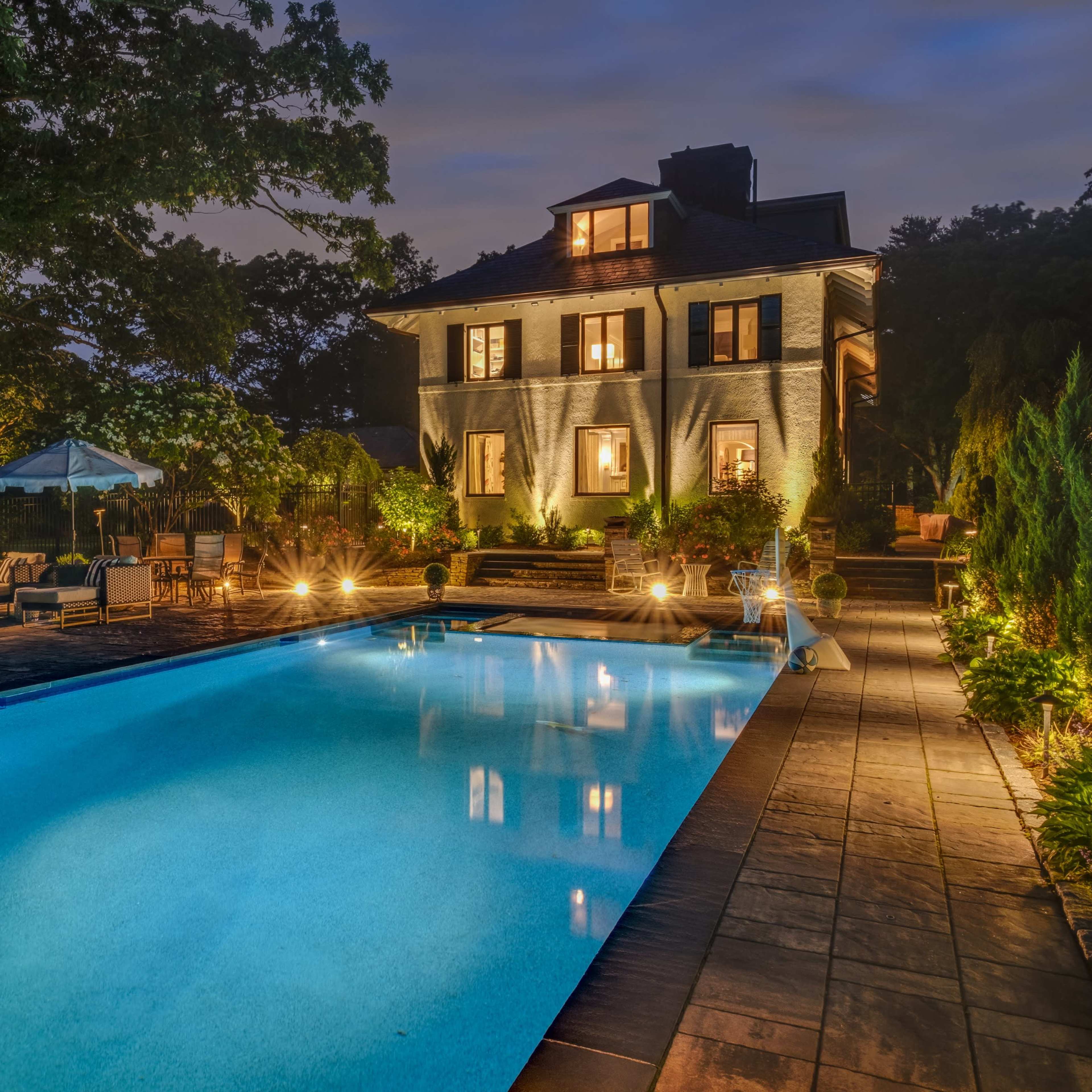A well-lit backyard features a swimming pool beside a two-story house with illuminated windows and landscaped greenery.