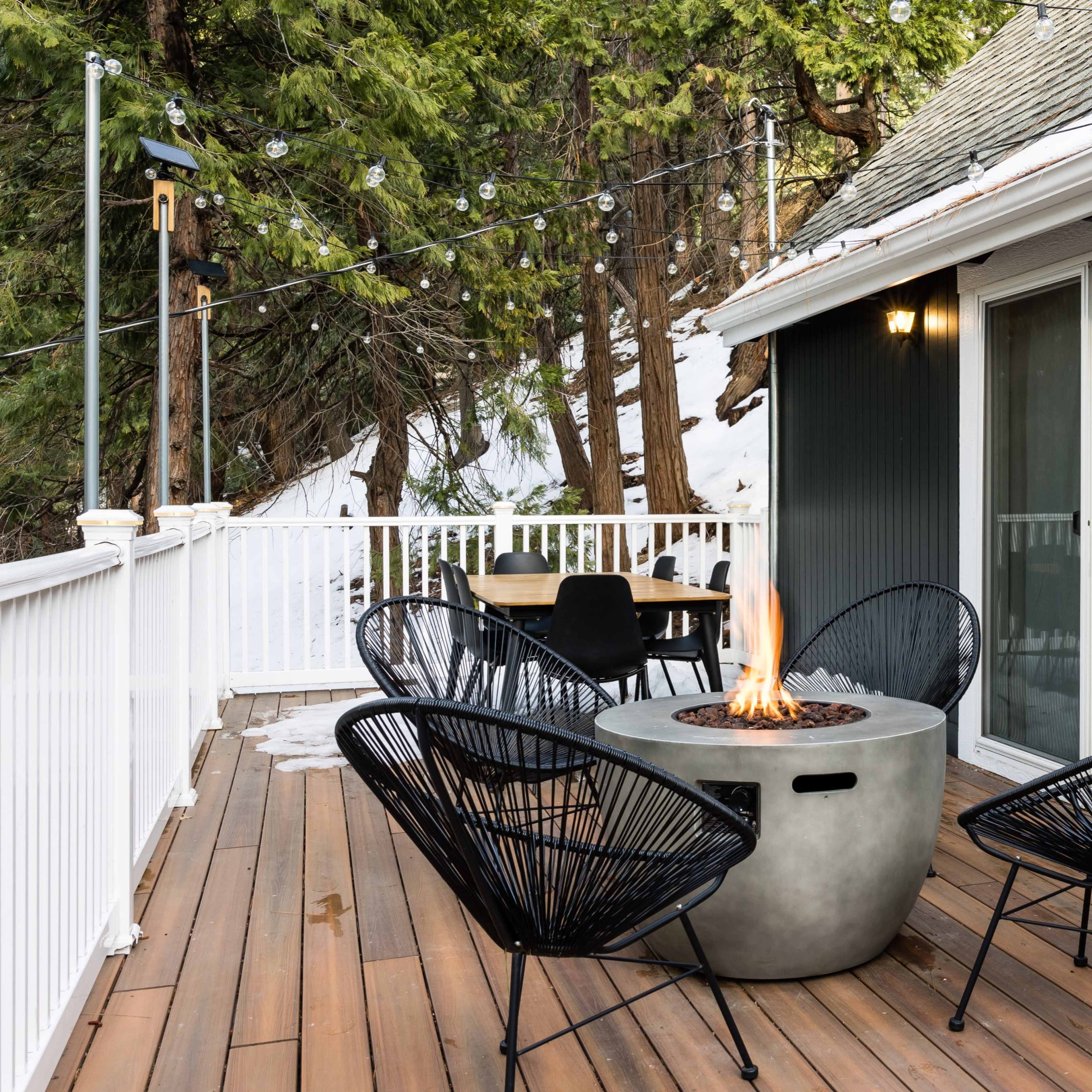 A modern deck features a circular fire pit surrounded by four chairs, with trees in the background and snow visible on the ground.