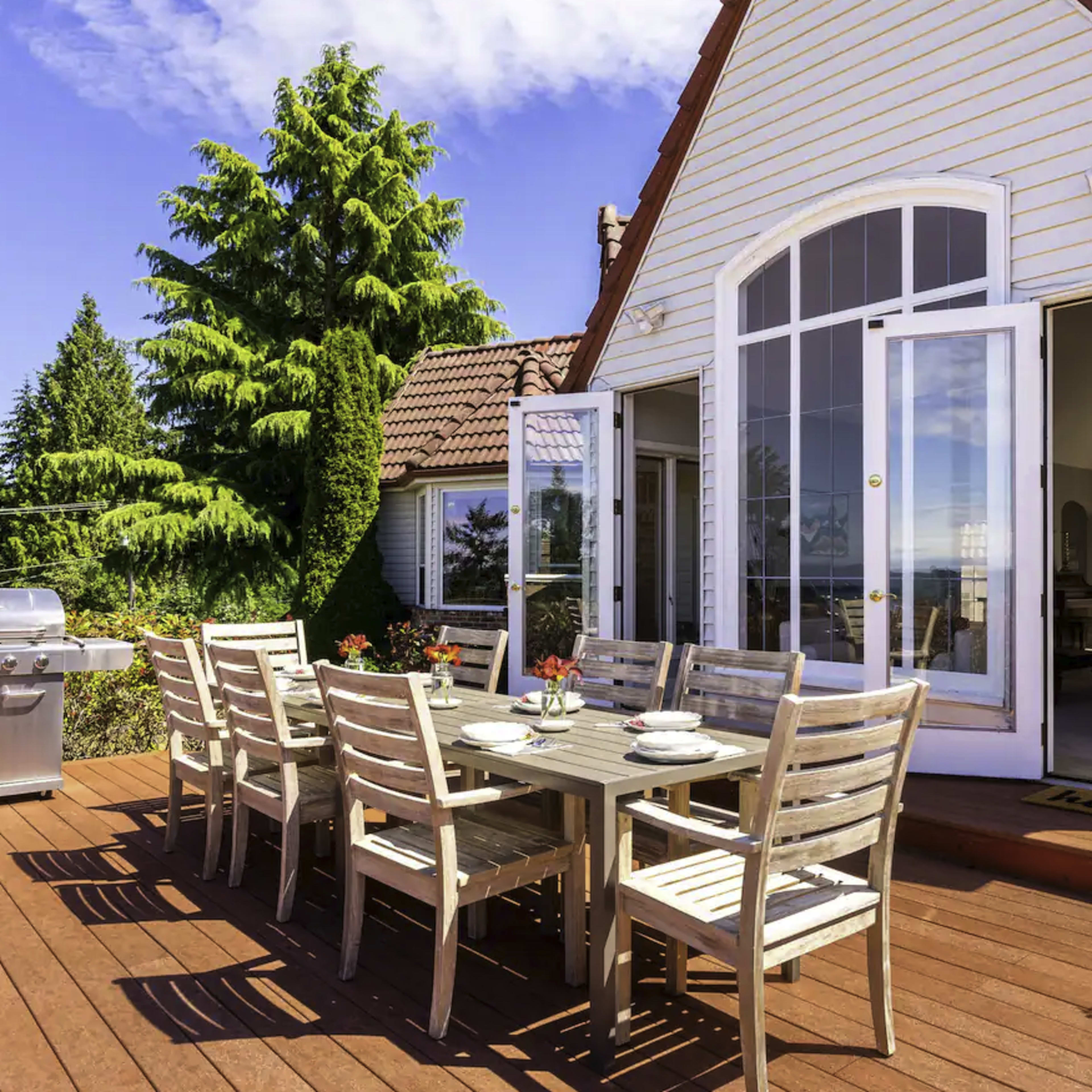 A wooden deck with a dining table and chairs is set up outside a house, featuring a grill and a view of trees and a blue sky.