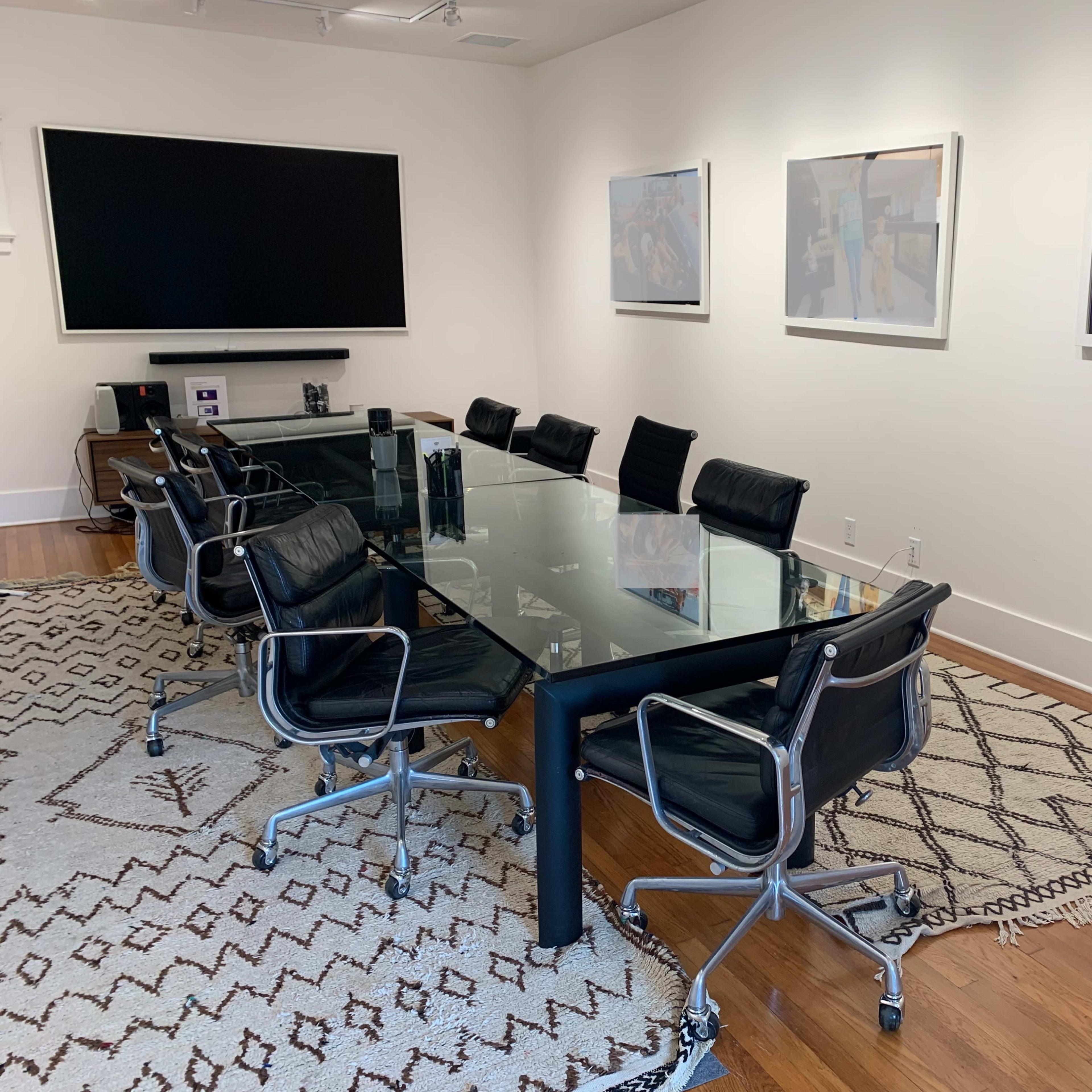 A modern conference room features a glass table surrounded by black chairs, with a large screen on one wall and artwork displayed on the opposite wall.