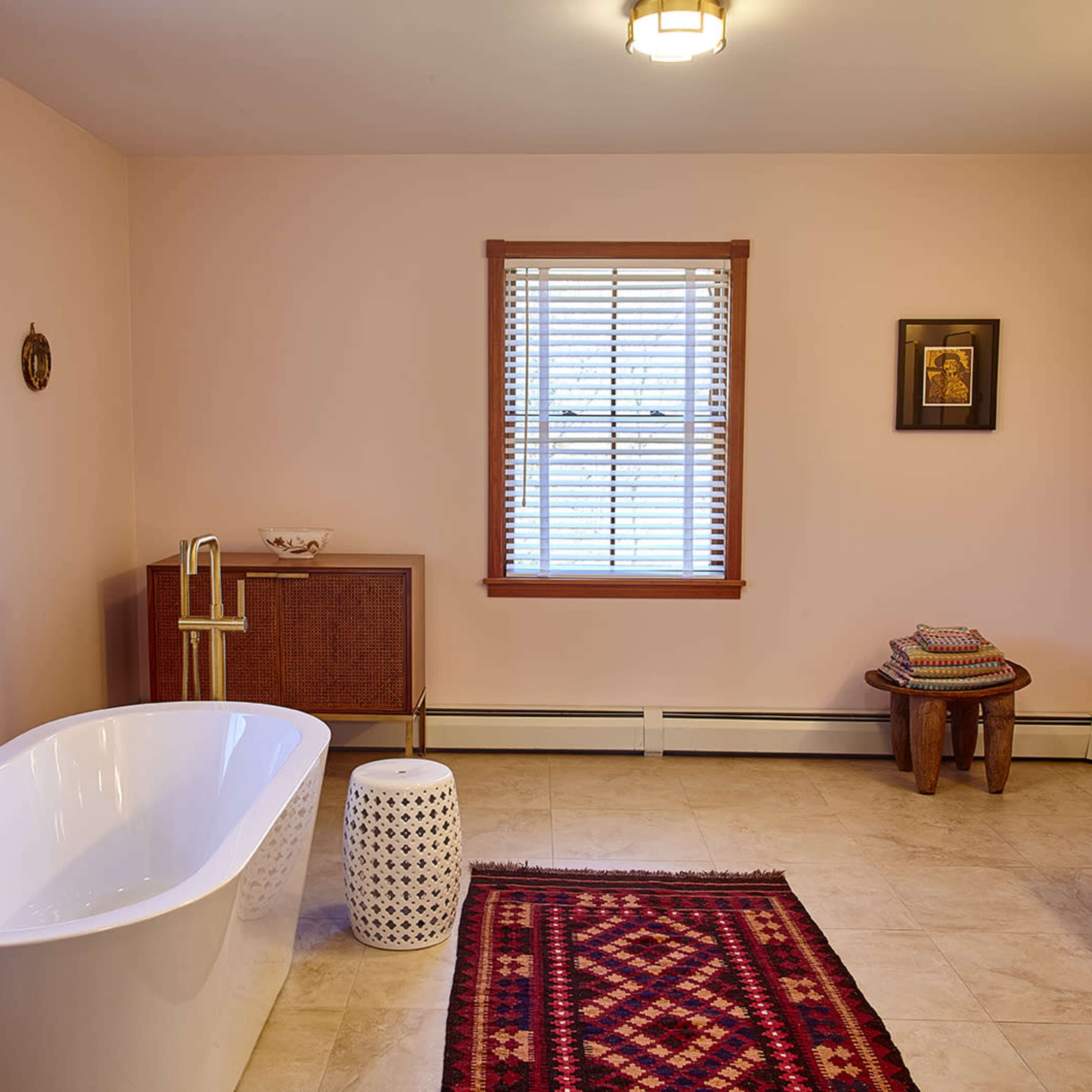 A bathroom featuring a freestanding bathtub, a window with blinds, a patterned rug, and a wooden cabinet.