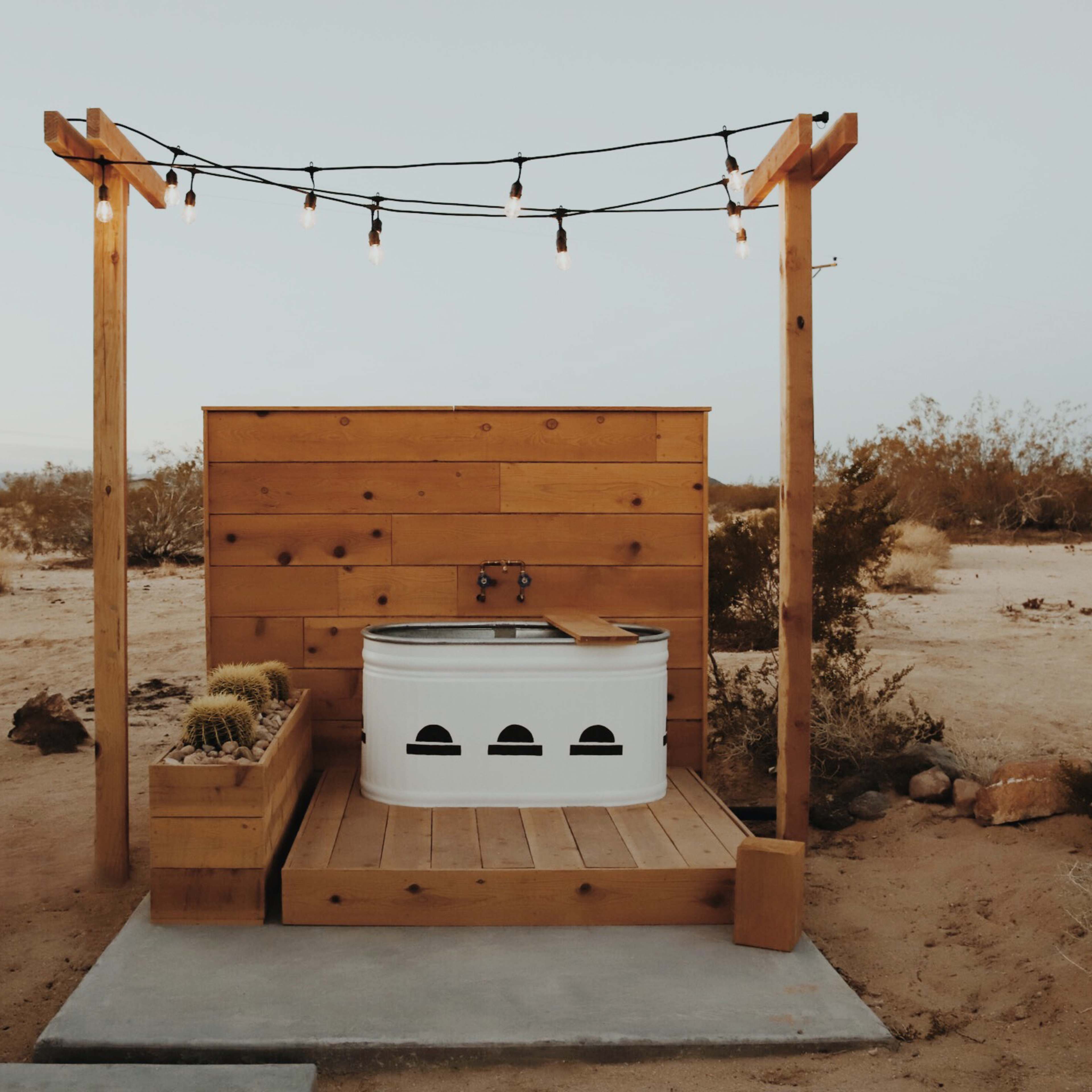 An outdoor hot tub enclosed by wooden decking and a rustic wooden wall, illuminated by string lights in a desert setting.