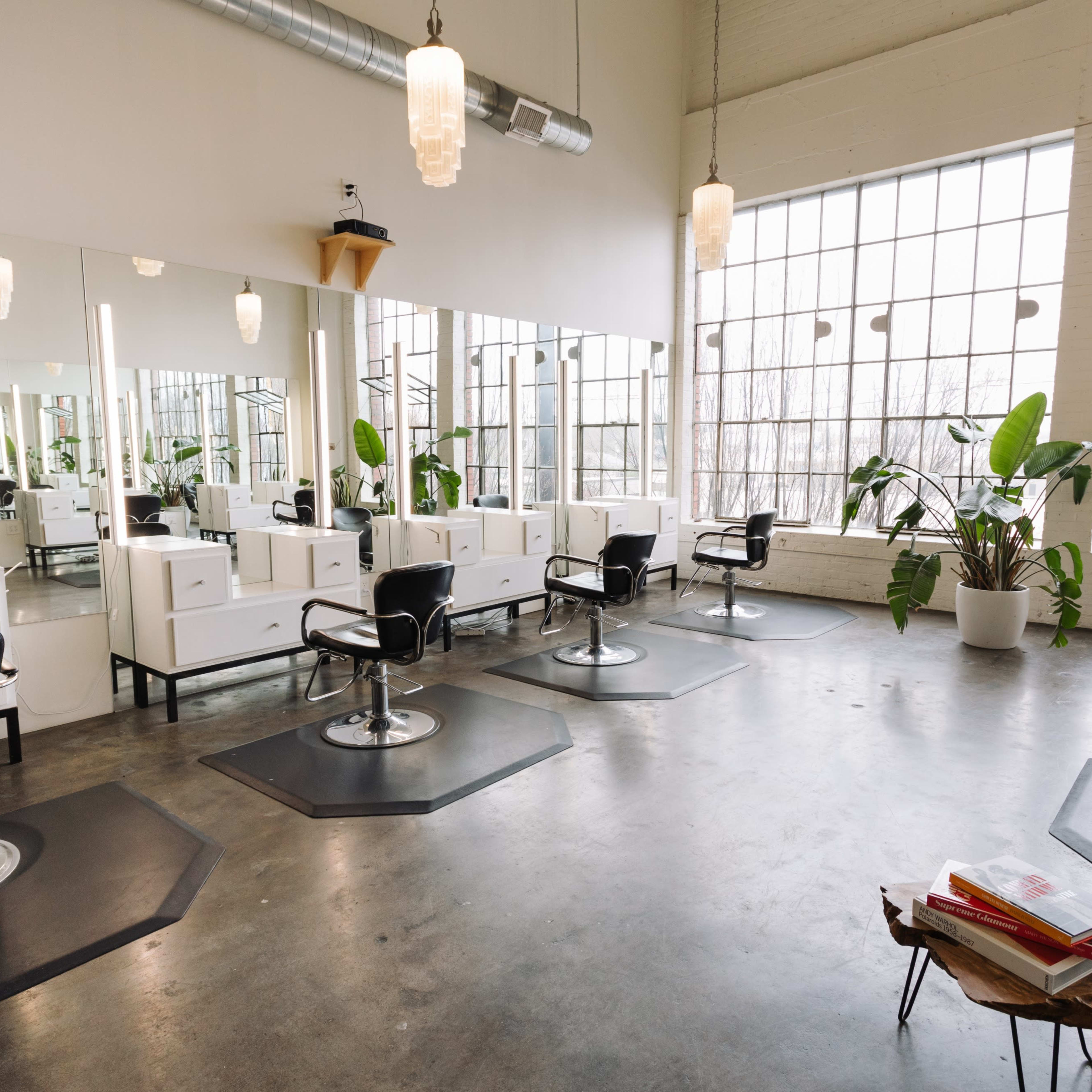 A spacious hair salon with several styling stations, large windows, and potted plants.