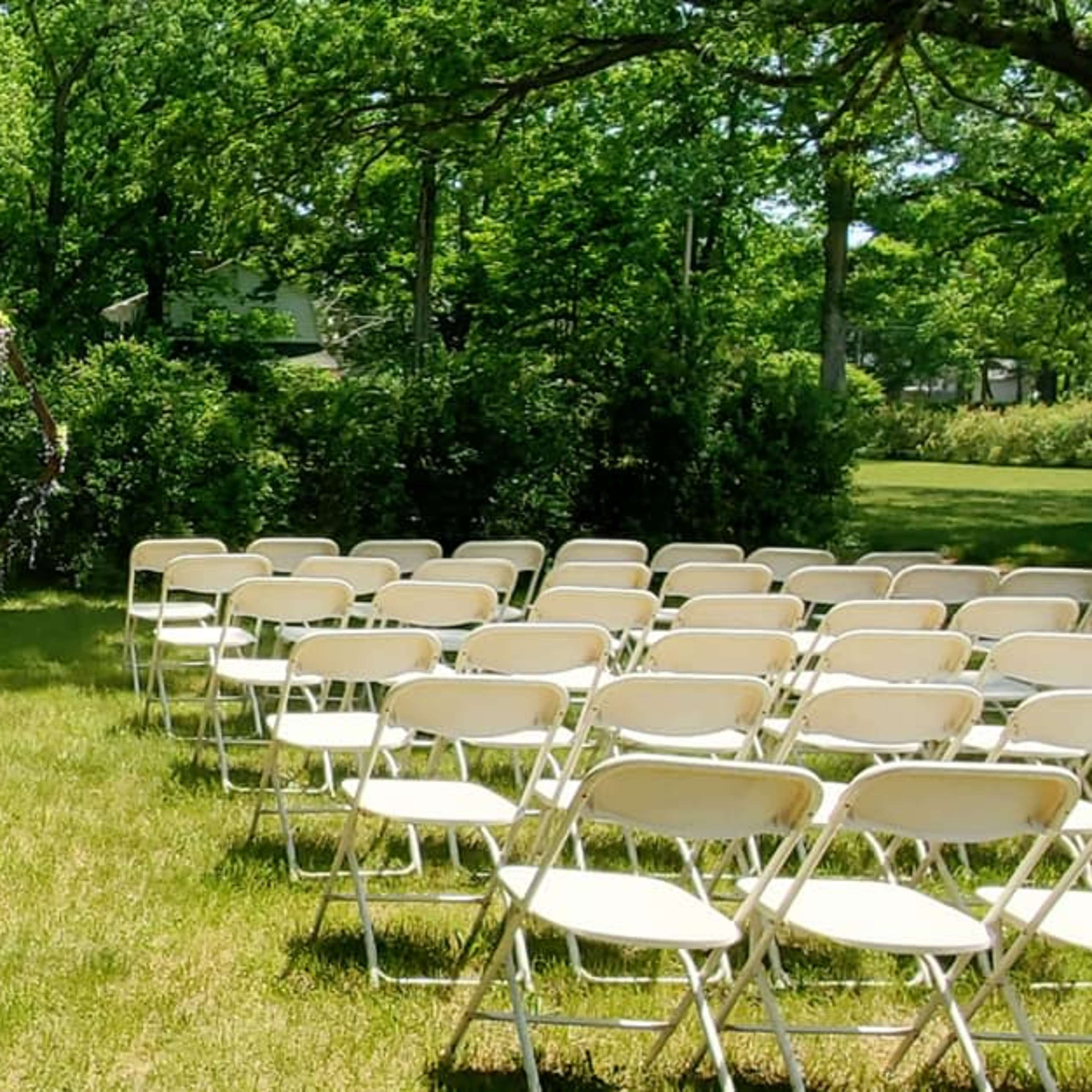 A grassy area is set up for an outdoor ceremony with rows of white folding chairs arranged facing a floral archway.