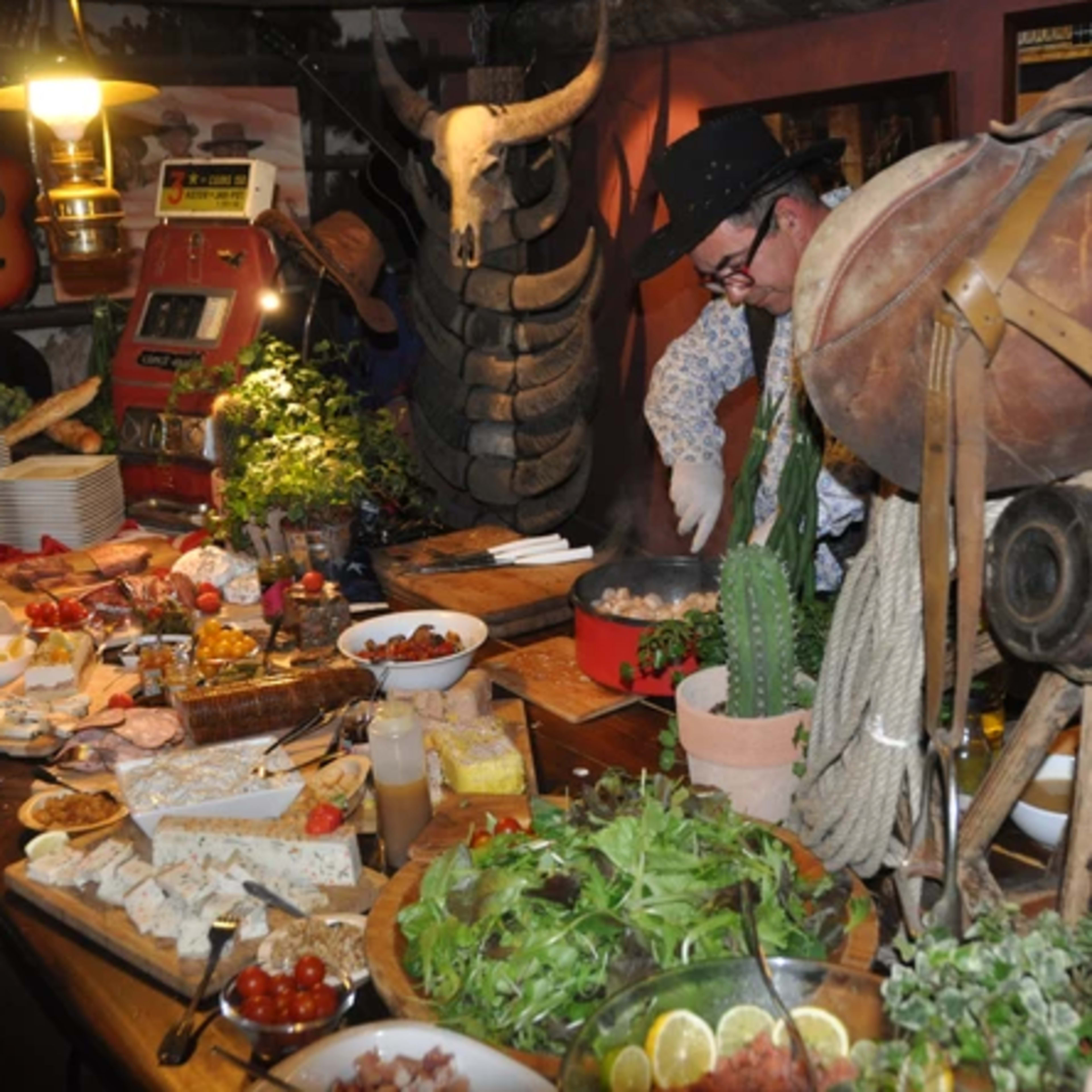 A rustic dining table is filled with various dishes, fresh ingredients, and cooking equipment, while a man in a cowboy hat prepares food nearby.
