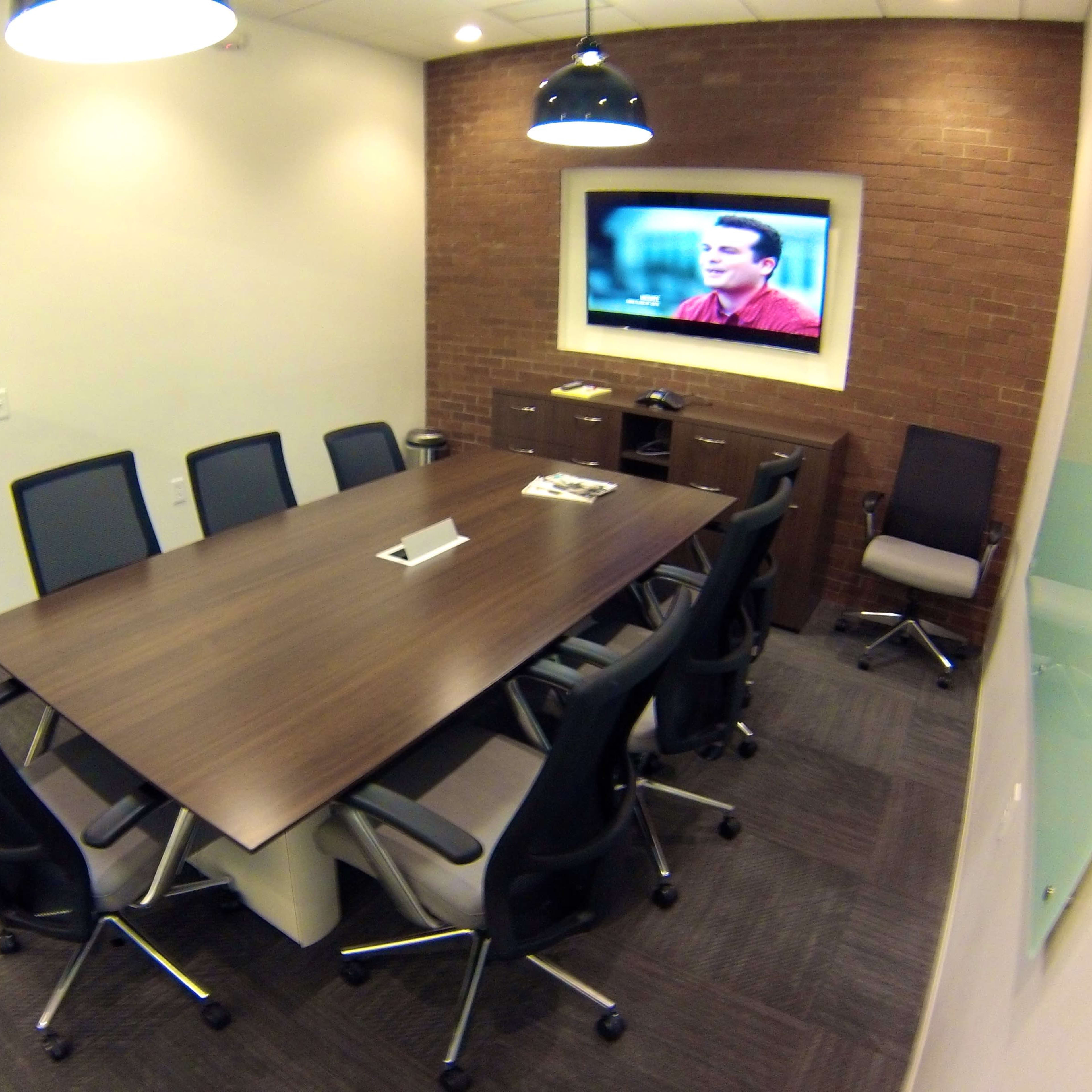 A conference room features a long wooden table surrounded by eight chairs, with a television mounted on the wall displaying a presentation.