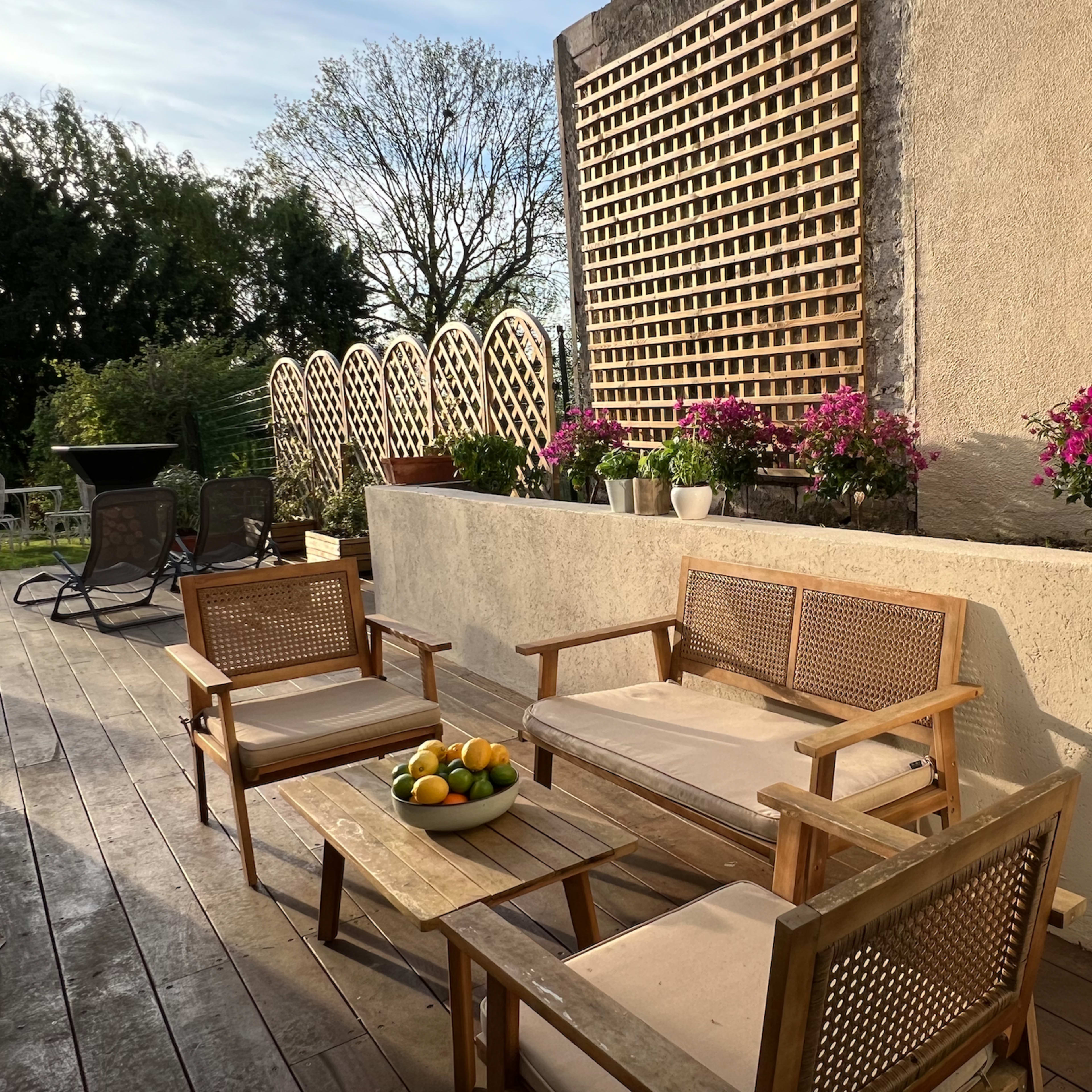 A wooden terrace features seating arrangements with a coffee table holding a bowl of lemons, surrounded by potted flowers and a decorative wall.