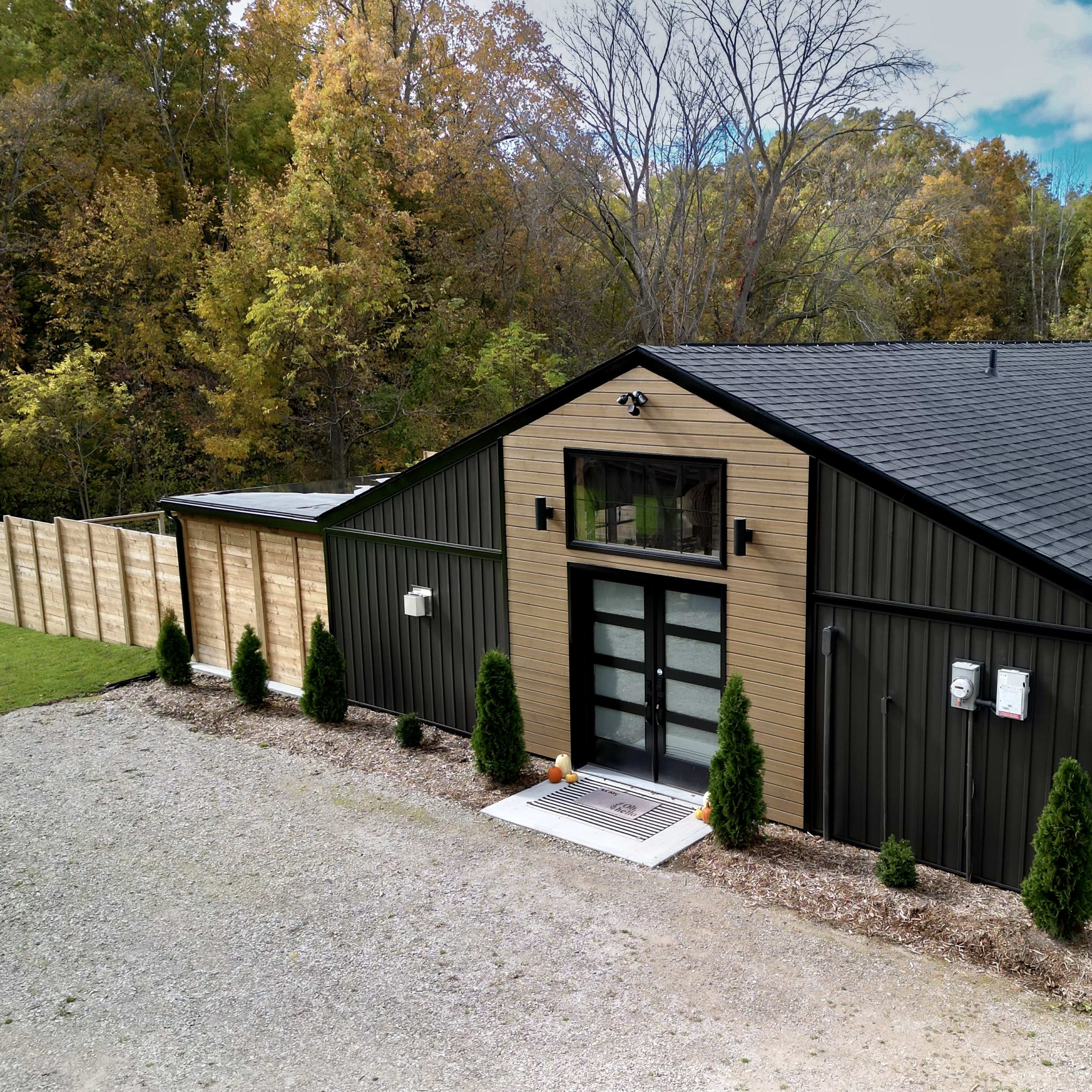 A modern, dark-colored building with a large glass entrance is surrounded by neatly trimmed shrubs and a gravel driveway, set against a backdrop of trees with autumn foliage.
