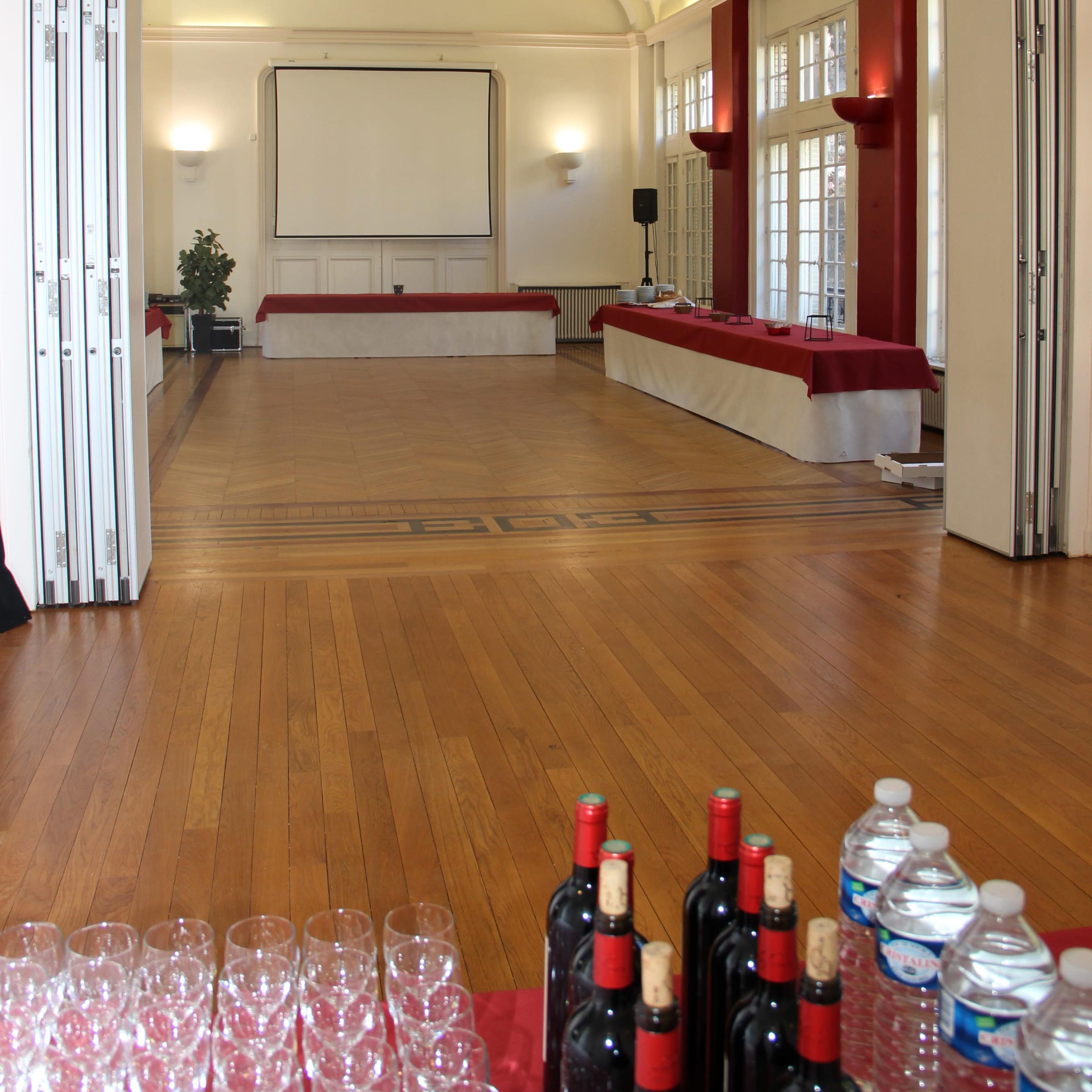 A spacious event room features a large empty stage area with a screen at the front and a table with red tablecloths along the back, accompanied by bottles of red wine and water on a nearby table.