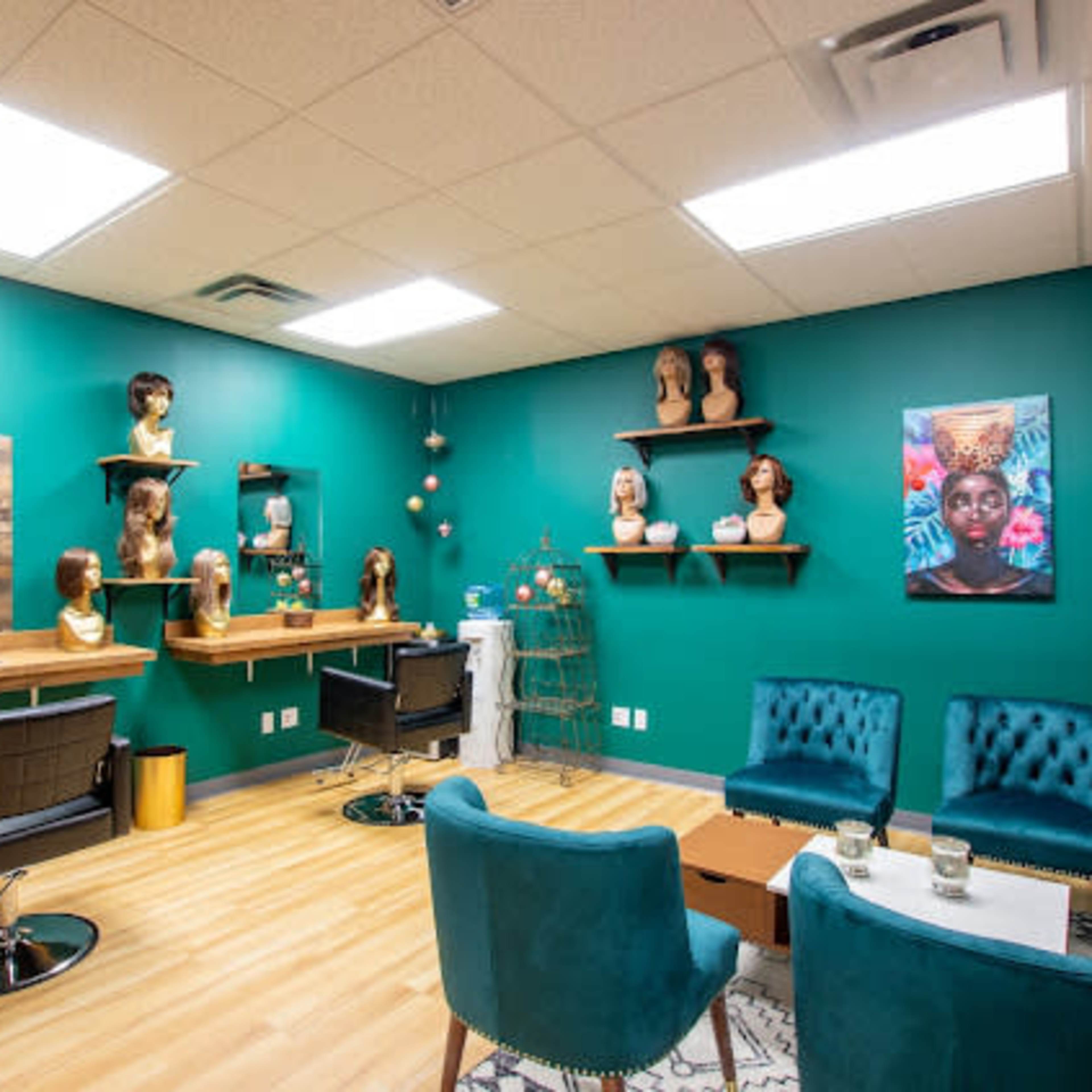 A hair salon interior featuring teal walls, a seating area with teal chairs, a wooden shelf displaying mannequin heads, and artwork on the wall.