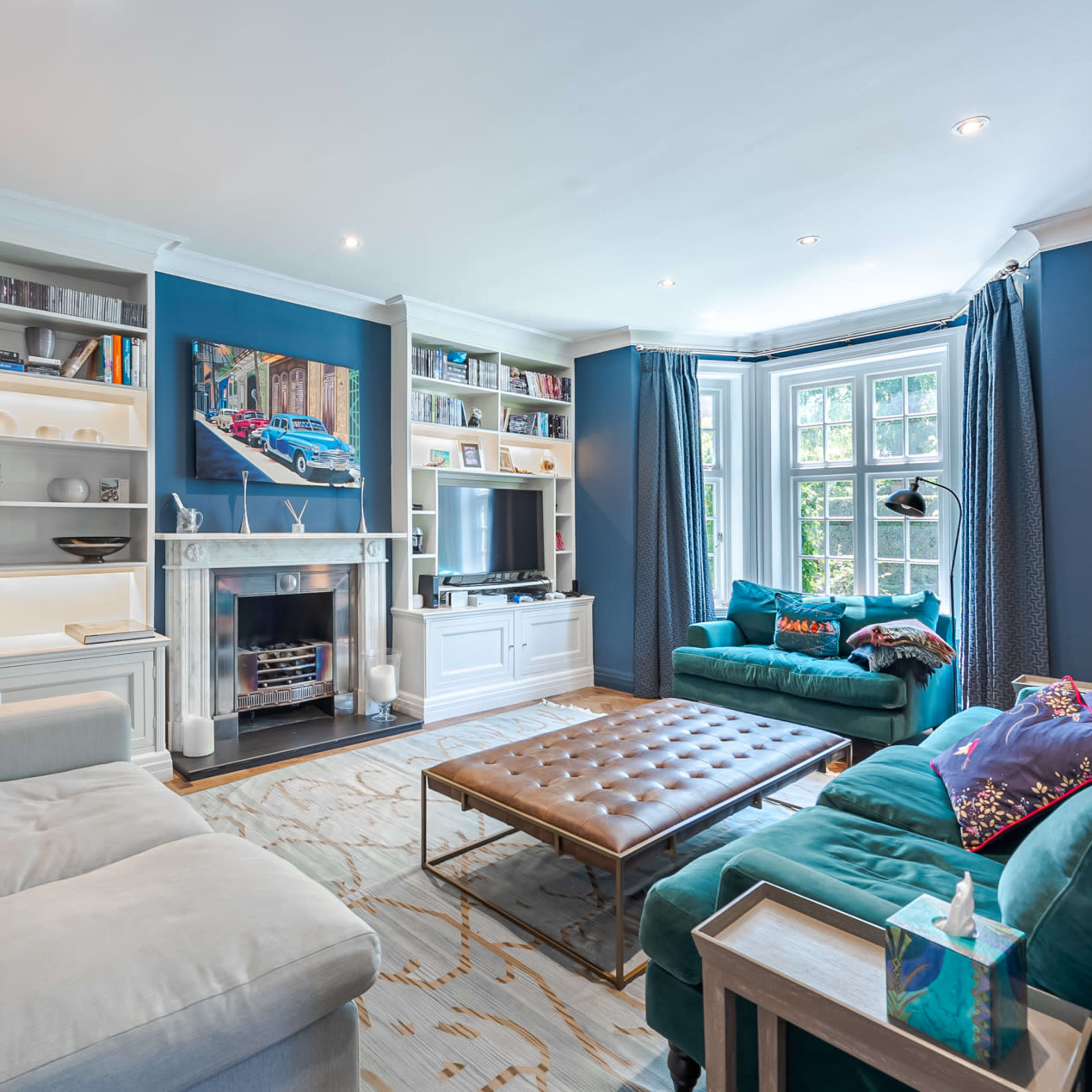 The living room features a blue color scheme with a sectional sofa, two teal armchairs, a bookshelf filled with books, and a large coffee table positioned in the center.
