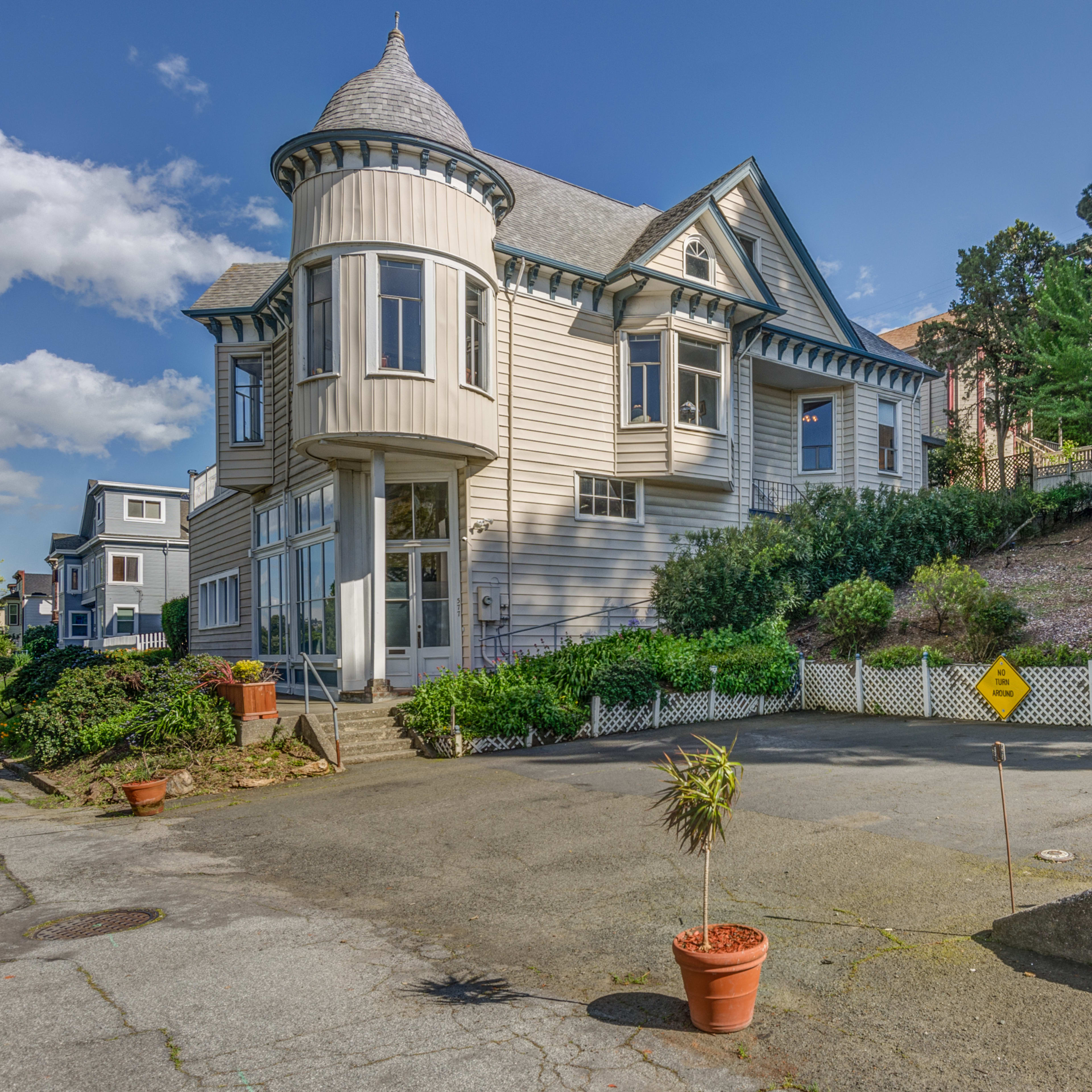 A Victorian-style house with a turret is situated on a sloped lot, surrounded by greenery and neighboring homes.