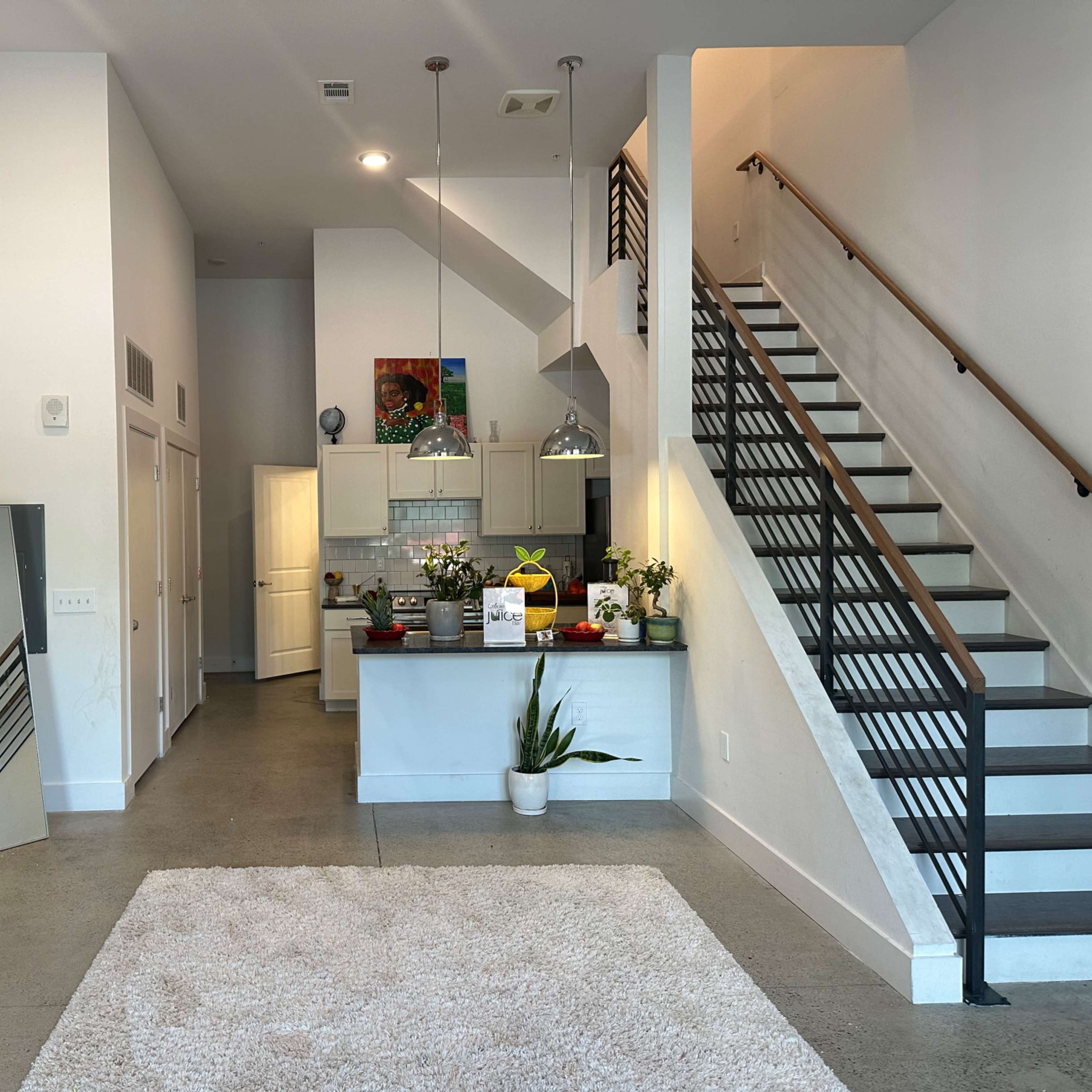 A modern foyer with a staircase, a small kitchen area, a large mirror, plants, and black folding chairs.