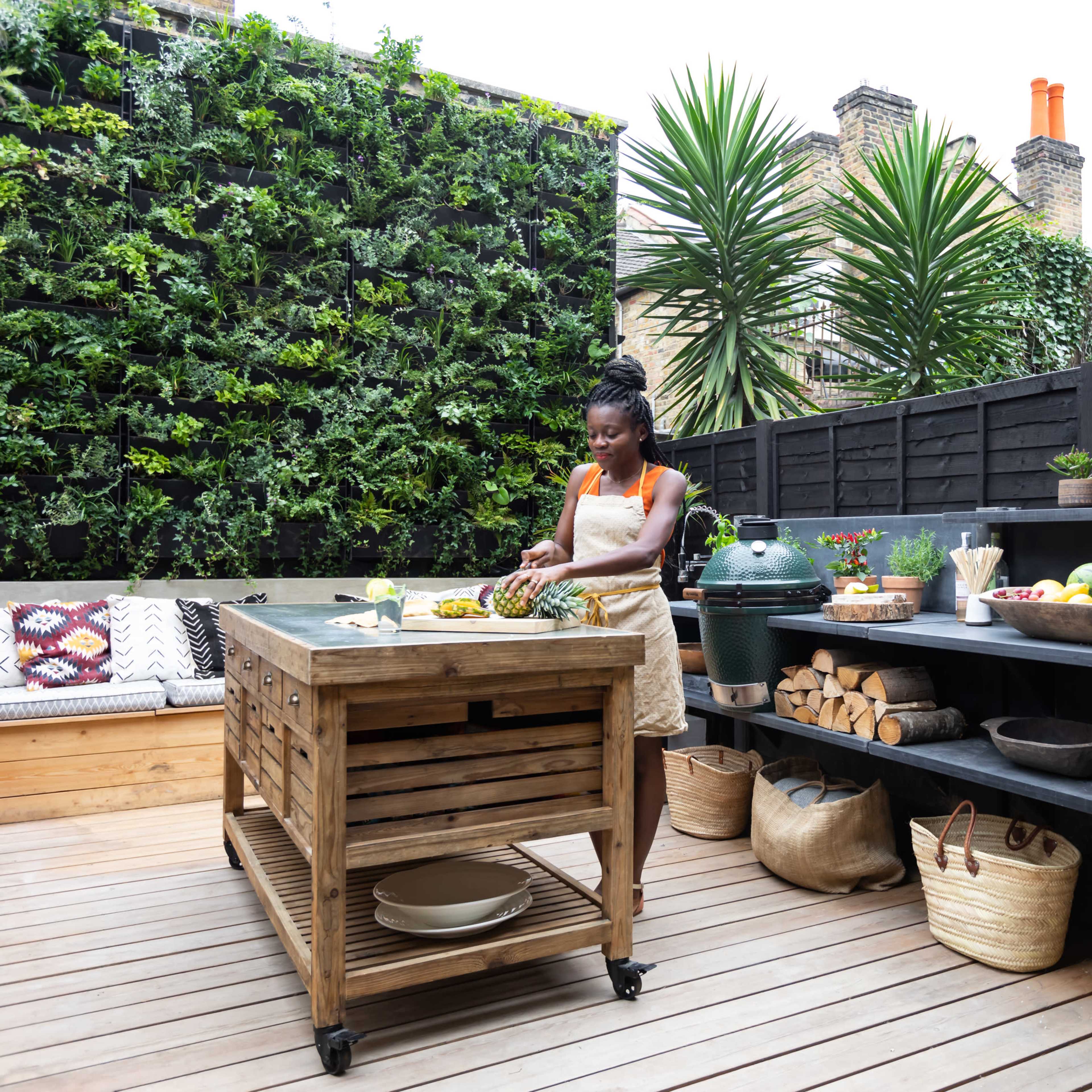 A woman prepares fruit on a wooden kitchen island in a modern outdoor kitchen surrounded by greenery.