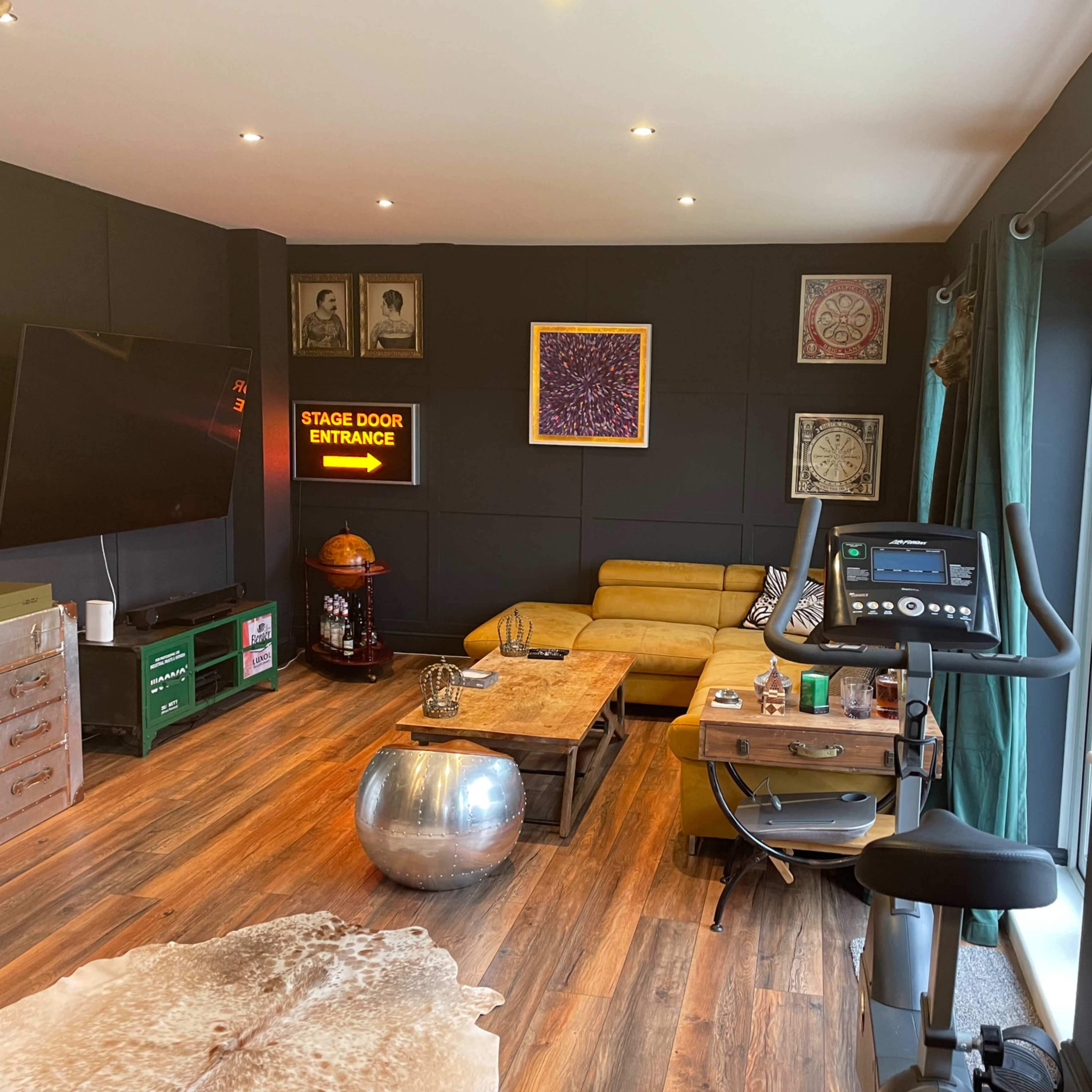 A modern room with a yellow sectional sofa, exercise bike, and a large television, complemented by various decor items and a wooden floor.
