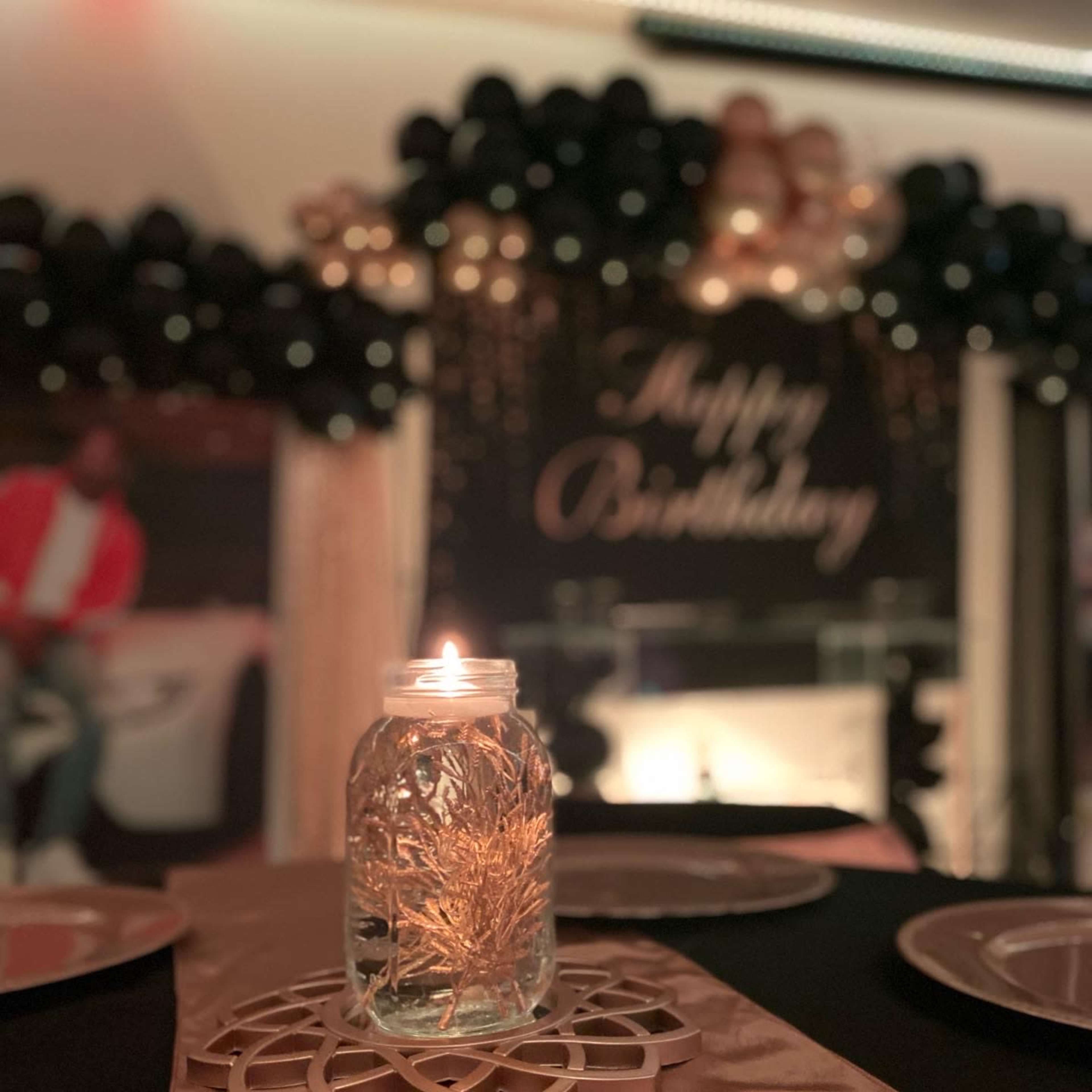A beautifully decorated table with a lit candle in a glass jar and a gold centerpiece, set against a backdrop of black balloons and a "Happy Birthday" sign.