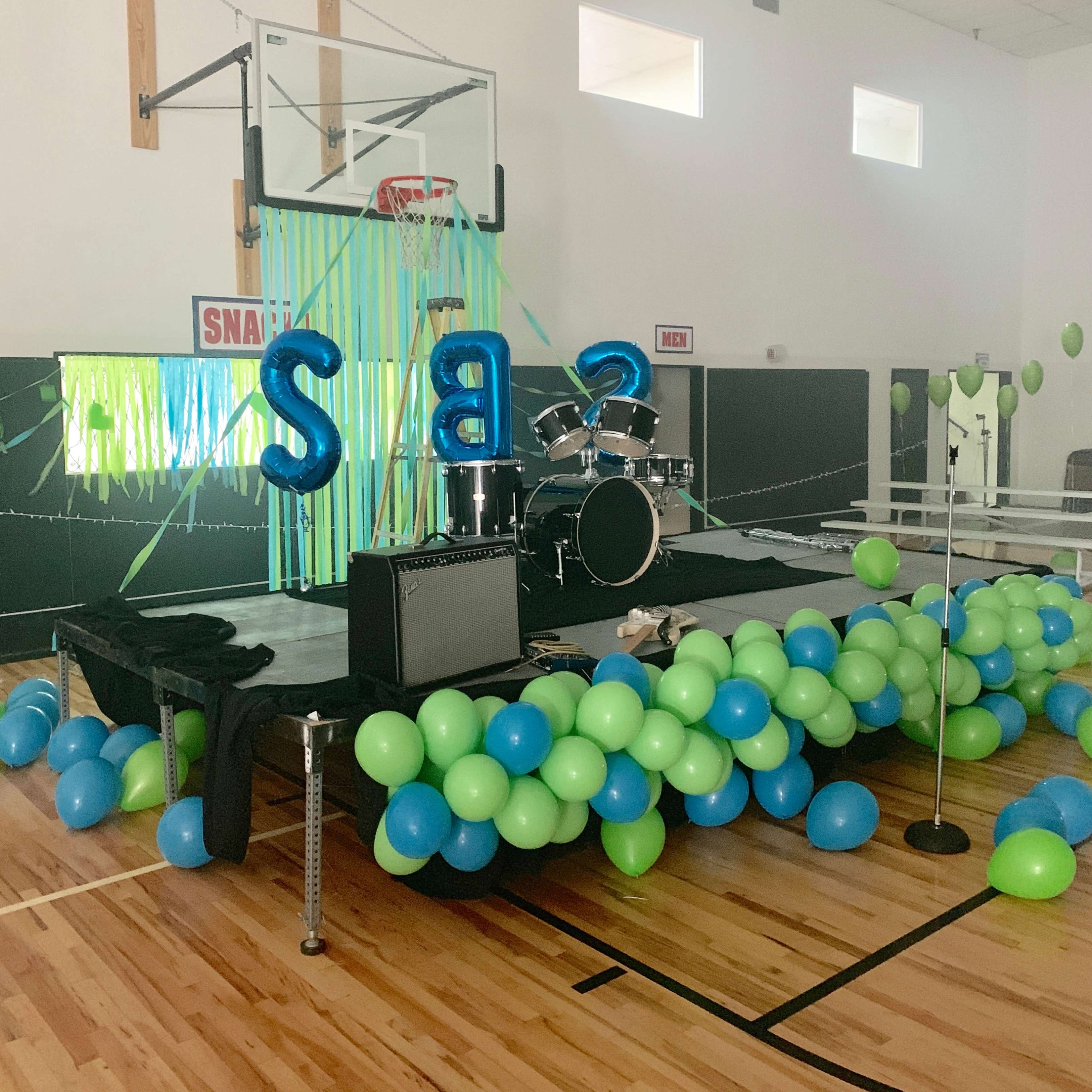 A decorated stage with a drum set and colorful balloons in shades of blue and green is set up in a gymnasium.