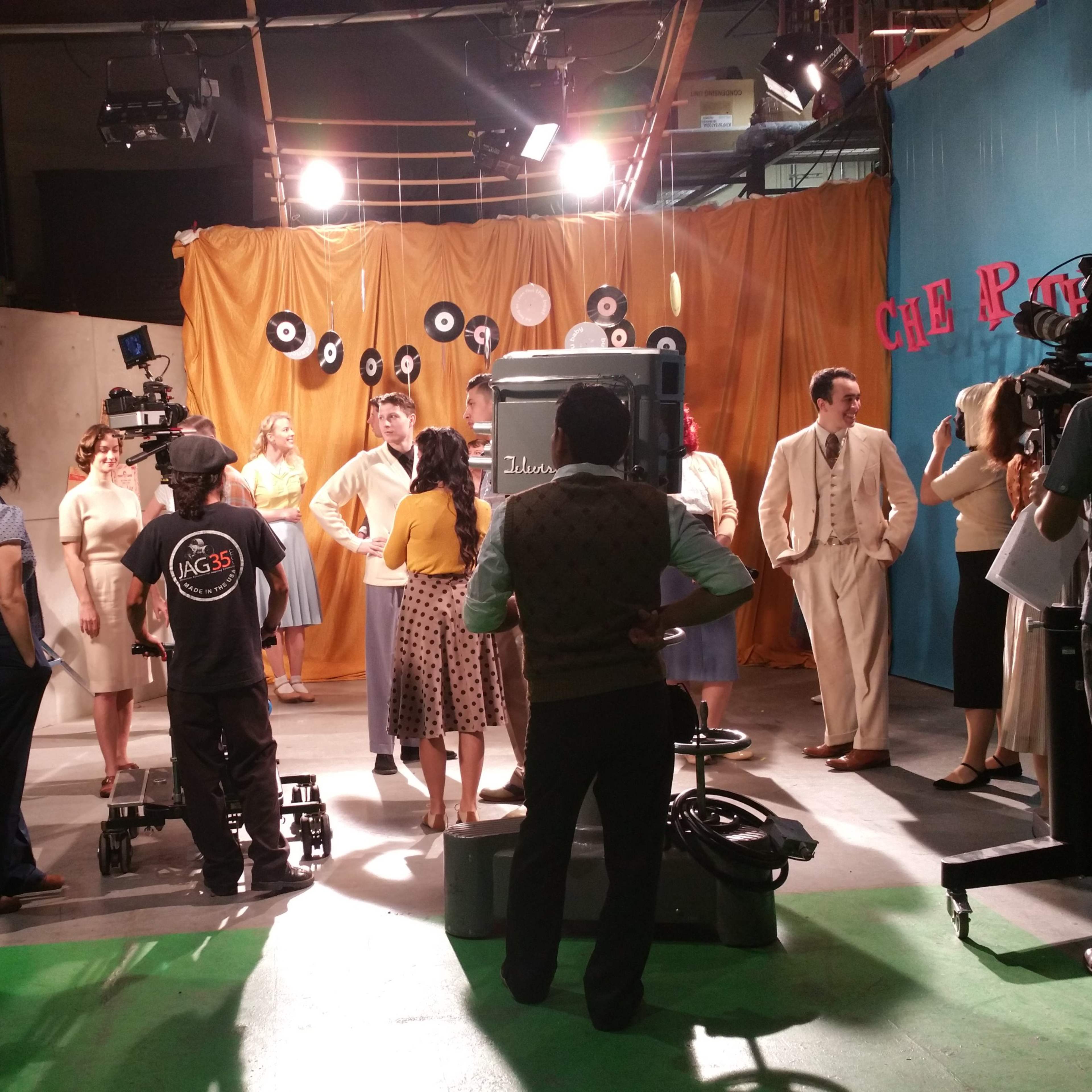 A cast and crew are gathered on a television set with vintage decor and equipment, preparing for a shoot.