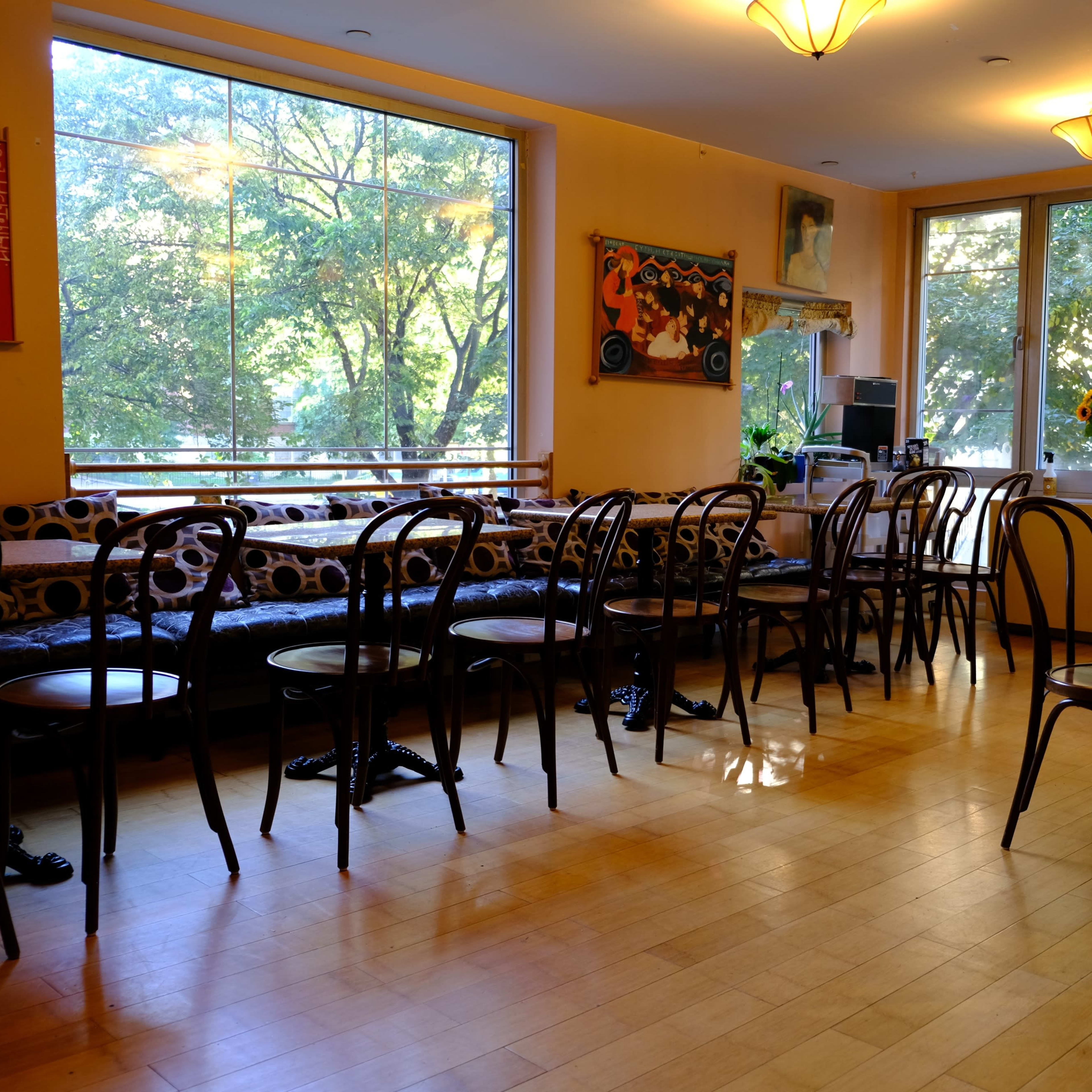 An empty café interior with wooden floors, several chairs arranged at tables, and large windows overlooking trees outside.