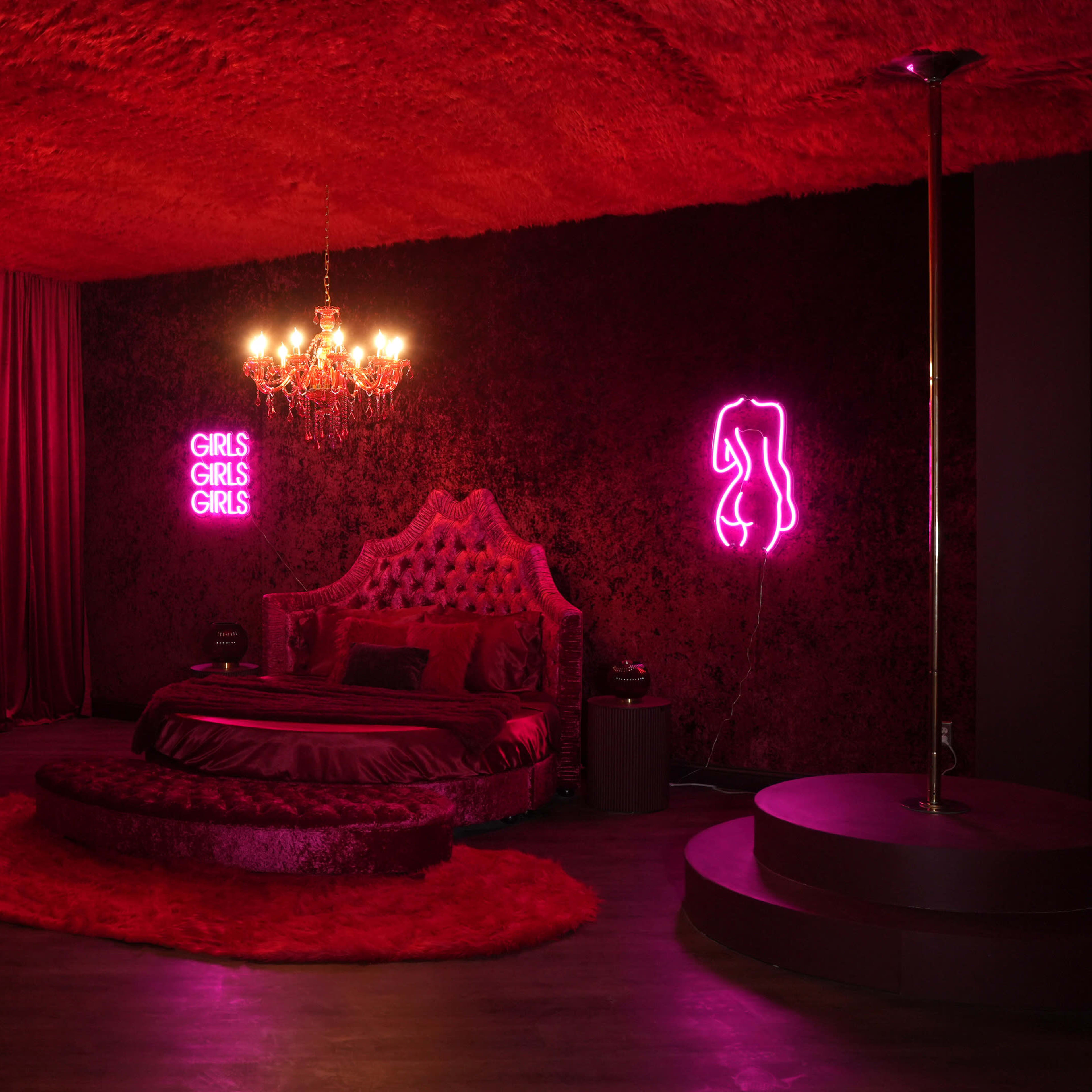 A richly decorated bedroom featuring red velvet walls, a large bed with ornate headboard, and neon signs that read "GIRLS GIRLS GIRLS."