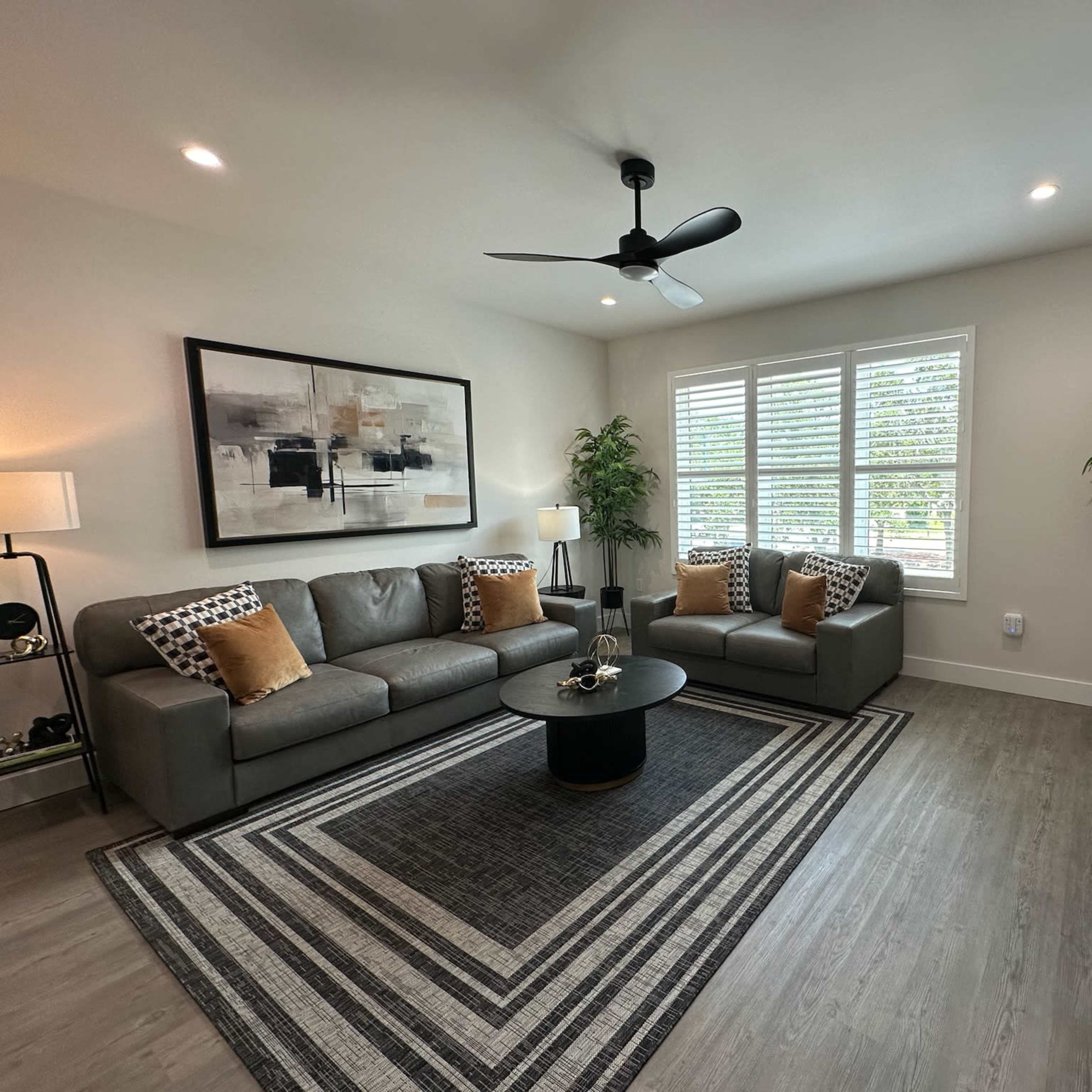 A modern living room with a gray sectional couch, a round black coffee table, and a large wall-mounted TV, surrounded by decorative plants and a geometric area rug.