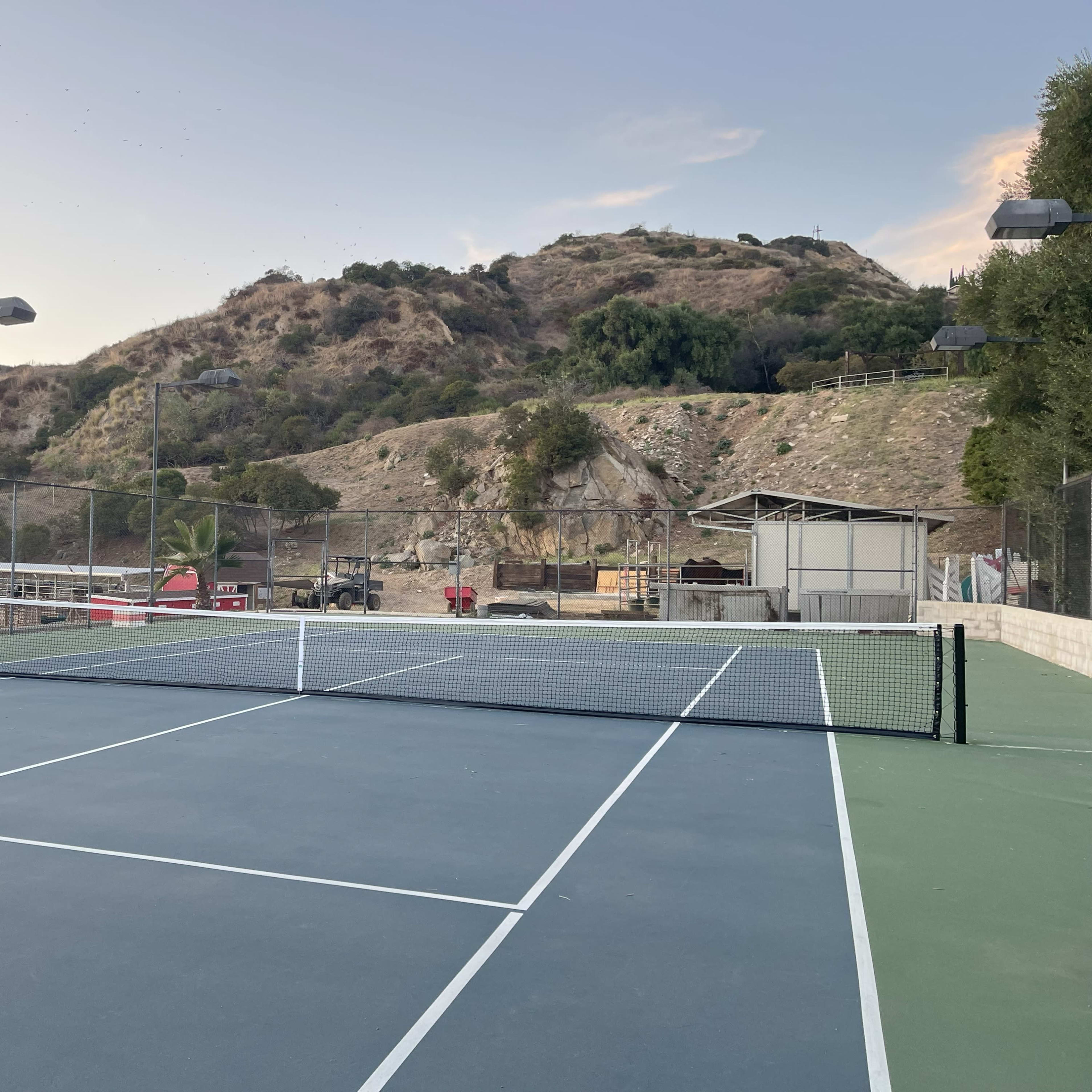 A tennis court is set against a backdrop of rolling hills and trees, with a nearby building and lights for evening play.
