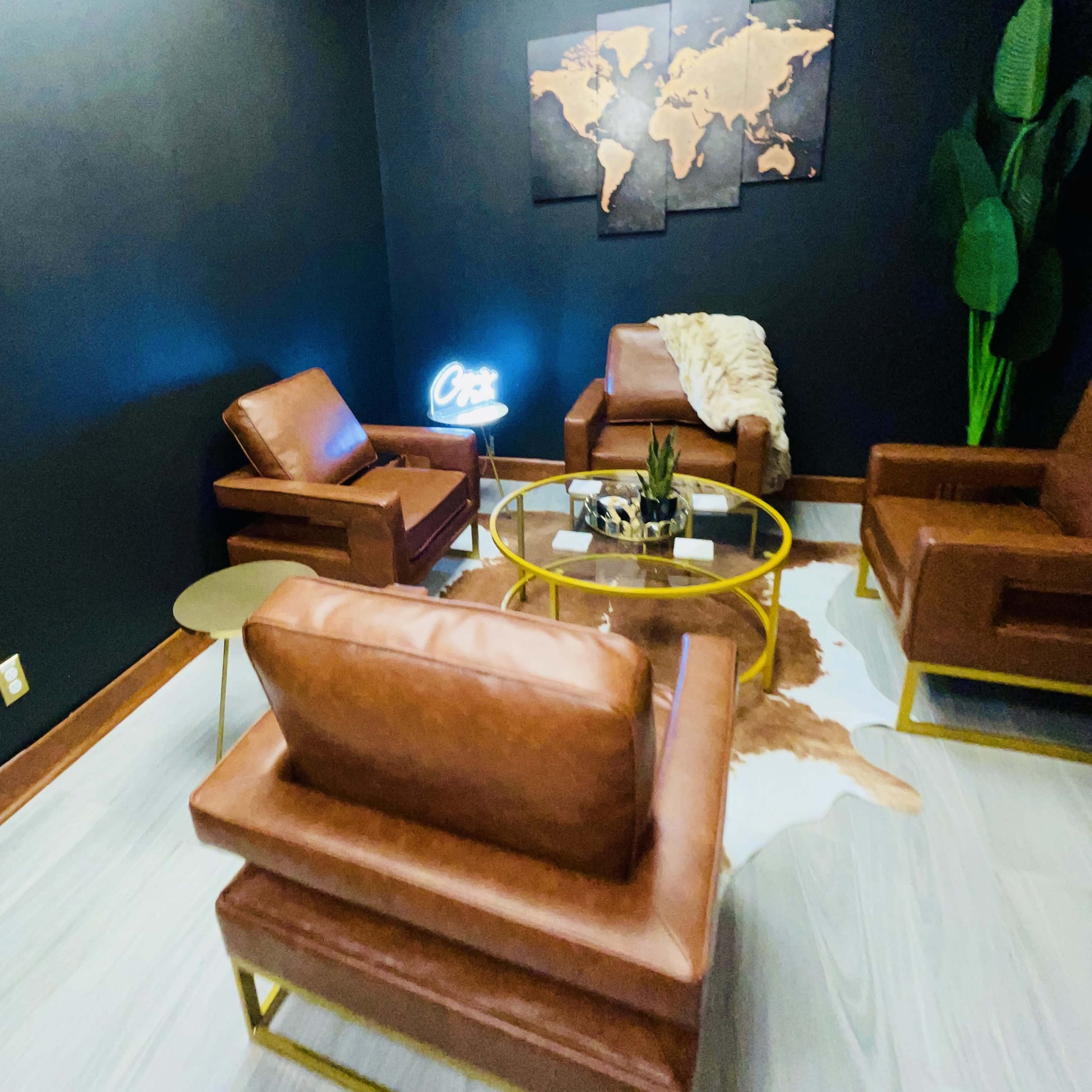 A small lounge area featuring two brown leather chairs, a round coffee table with decorative items, a cowhide rug, and a world map on the wall, all complemented by a neon sign.