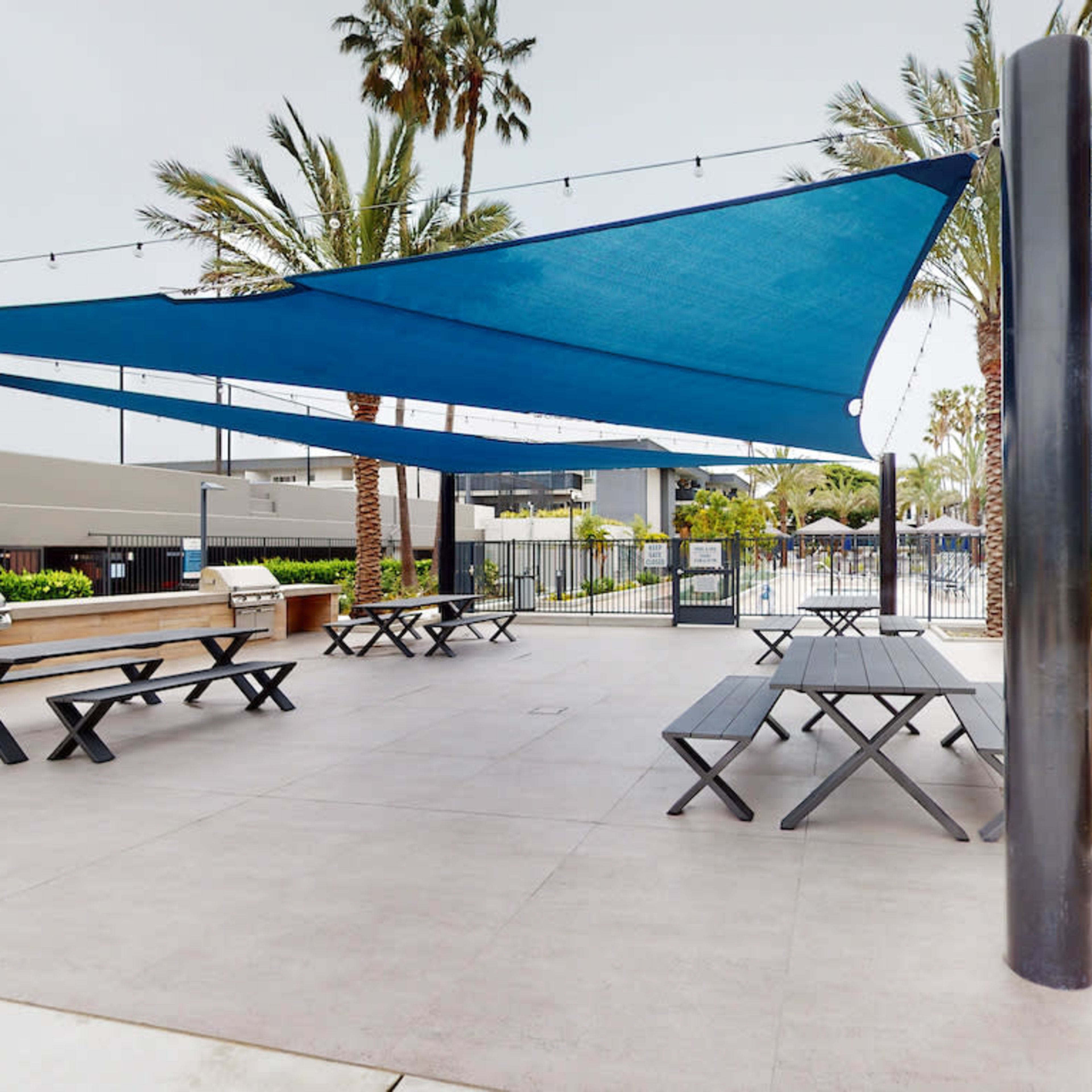 An outdoor barbecue area with several picnic tables shaded by blue canopies, surrounded by palm trees.