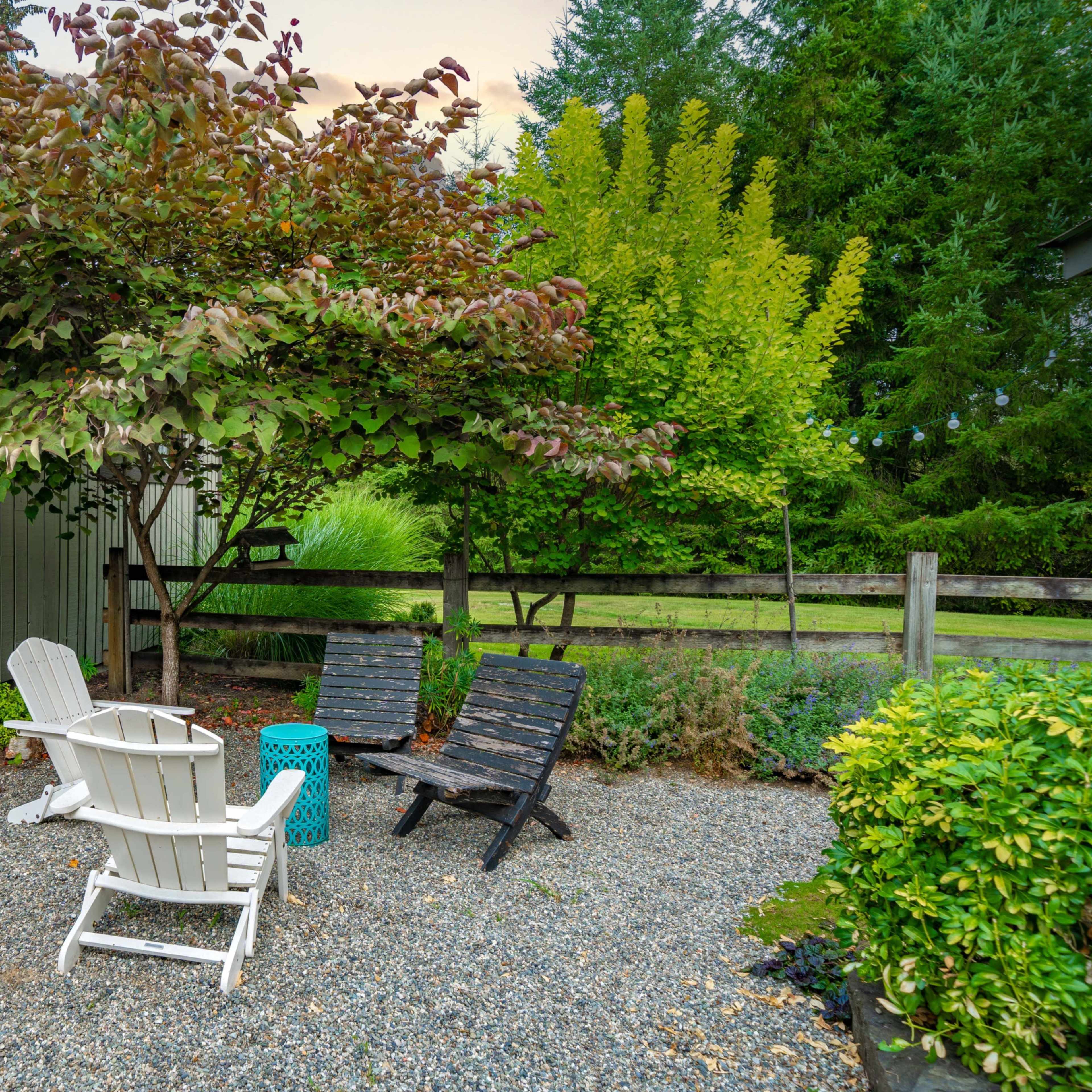 A gravel area features two chairs and a small table beneath a tree, surrounded by lush greenery.