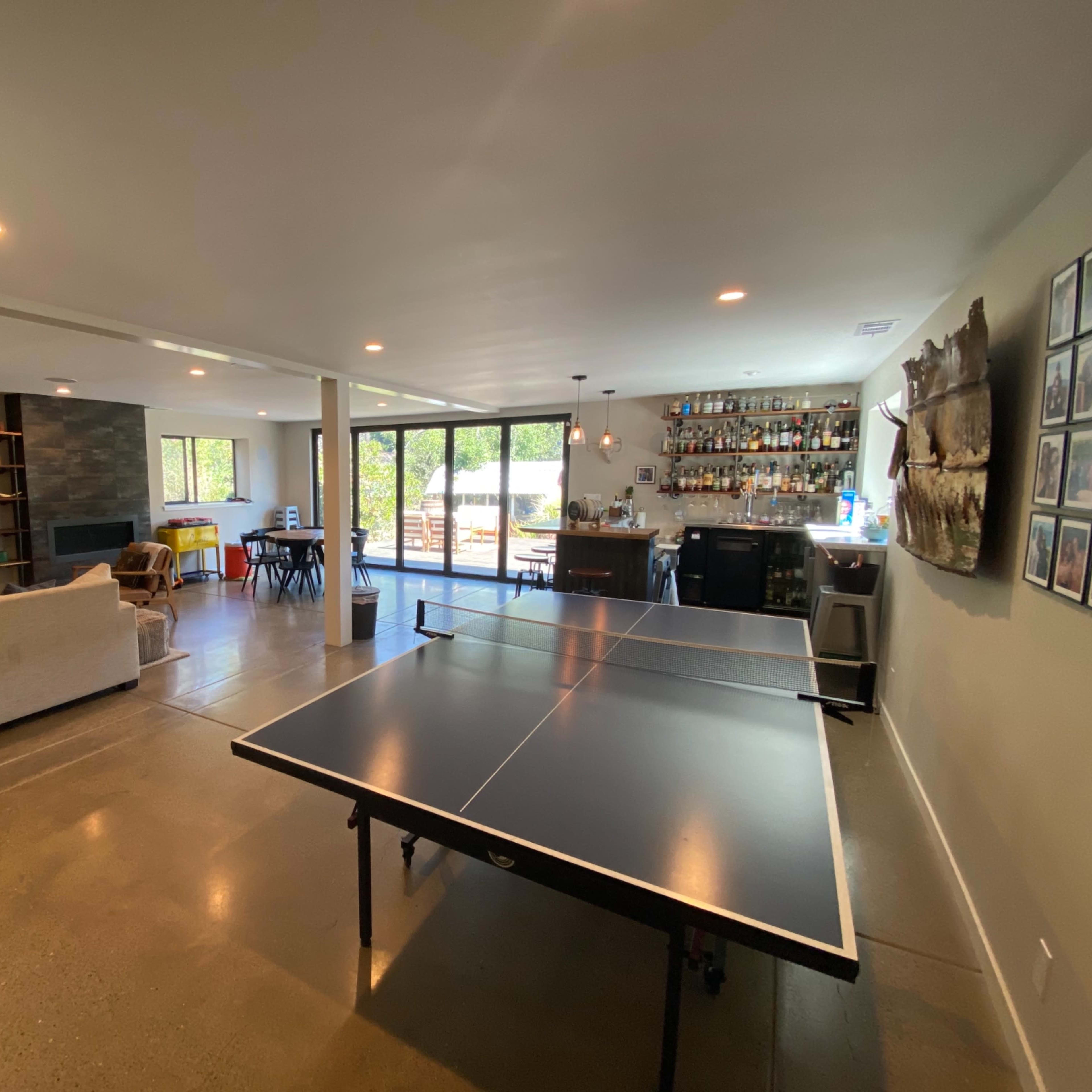 A spacious room featuring a ping pong table, a bar area with shelves of bottles, and large windows overlooking an outdoor space.