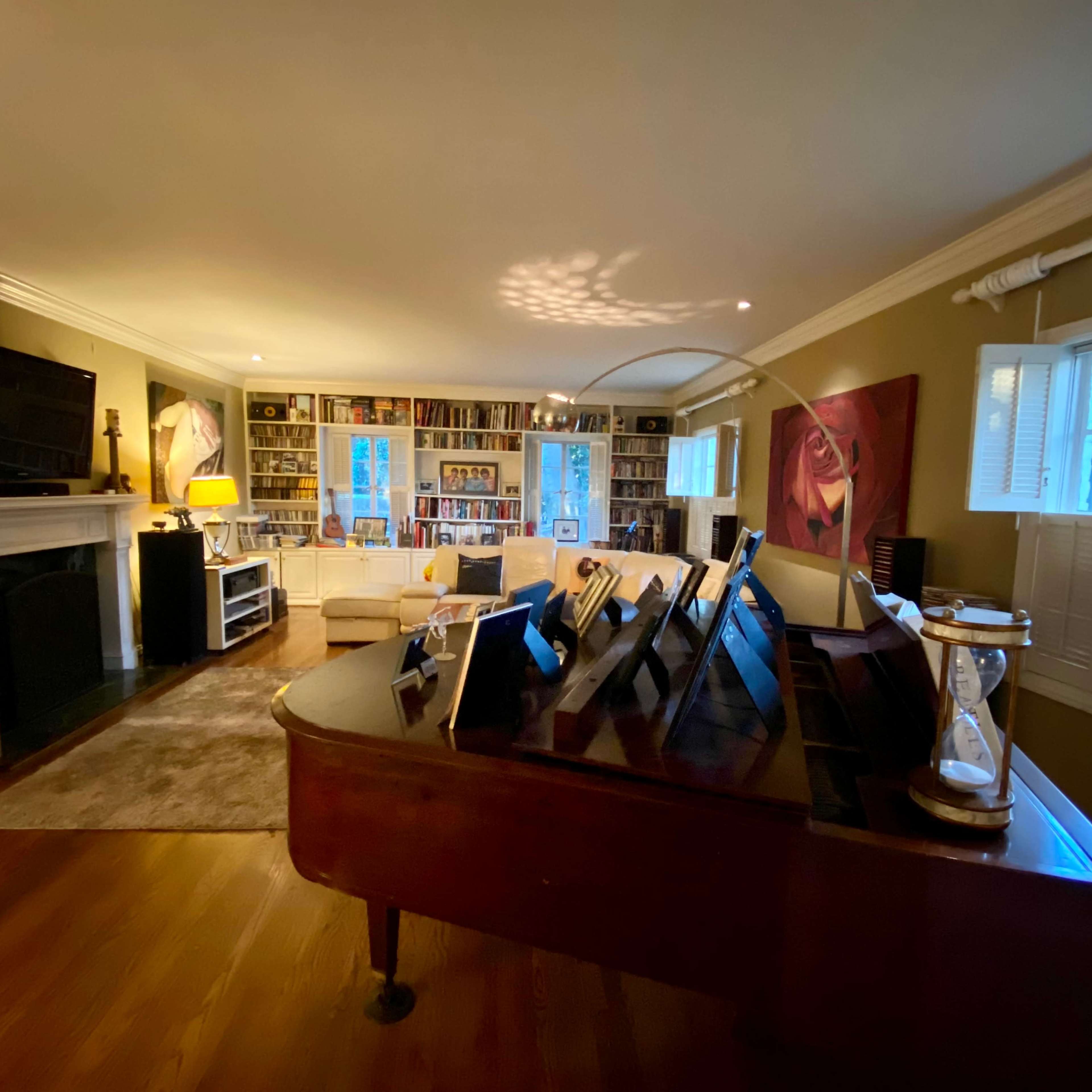 A living room features a grand piano in the foreground, with bookshelves and a comfortable seating area visible in the background.