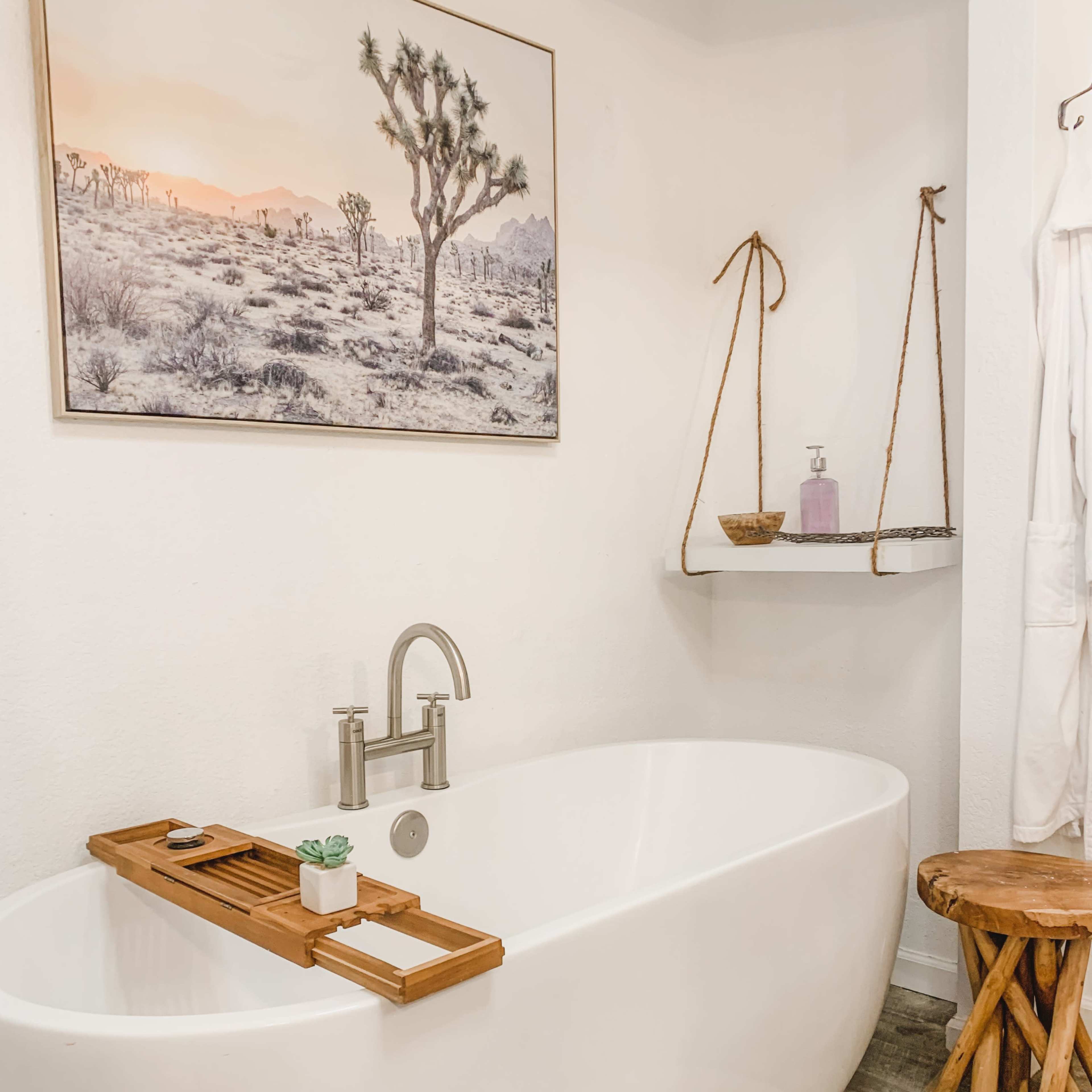 A modern bathroom featuring a freestanding bathtub, a wall-mounted shelf with decorative items, and a desert landscape picture above the tub.