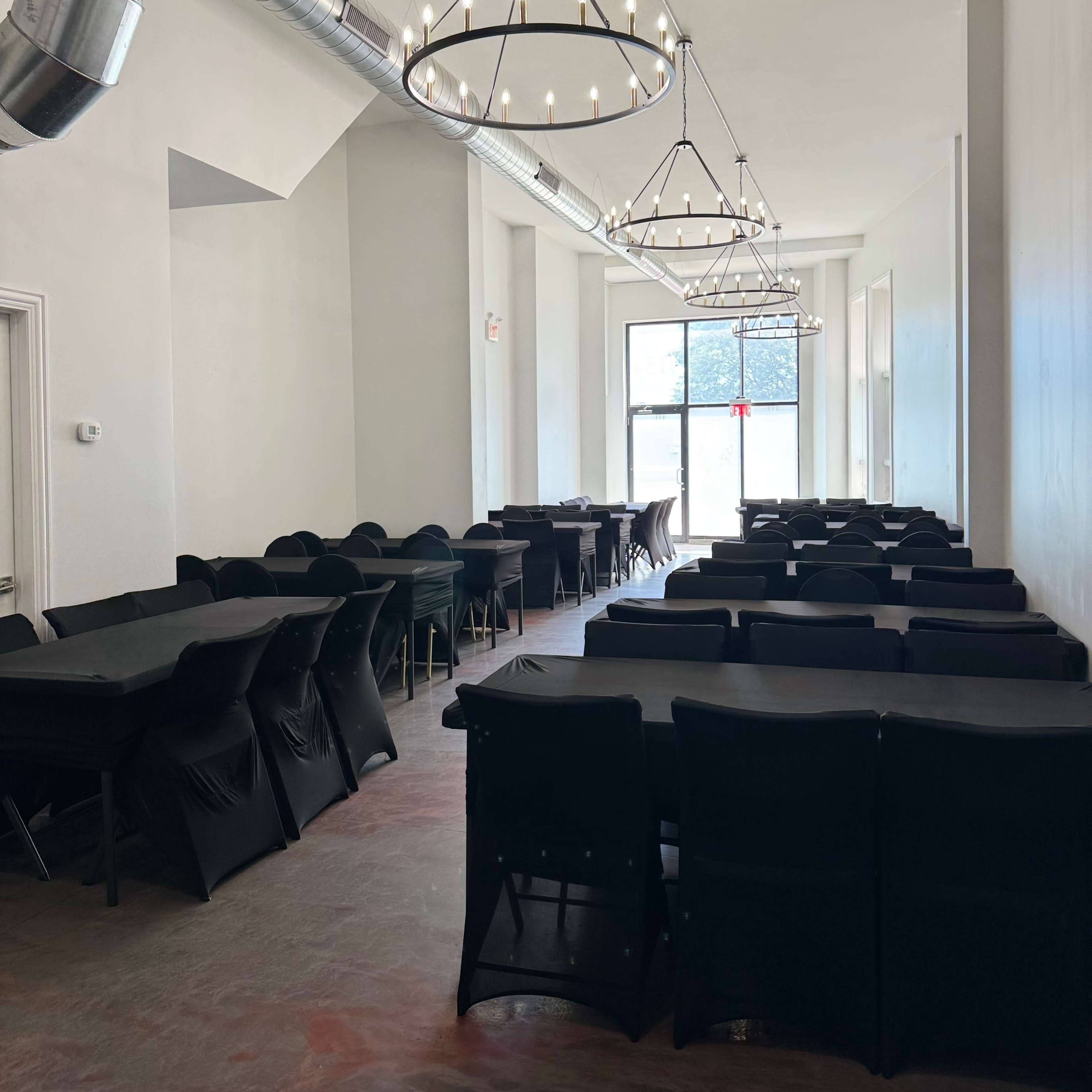 A long, narrow dining area with black tables and chairs under industrial-style chandeliers.