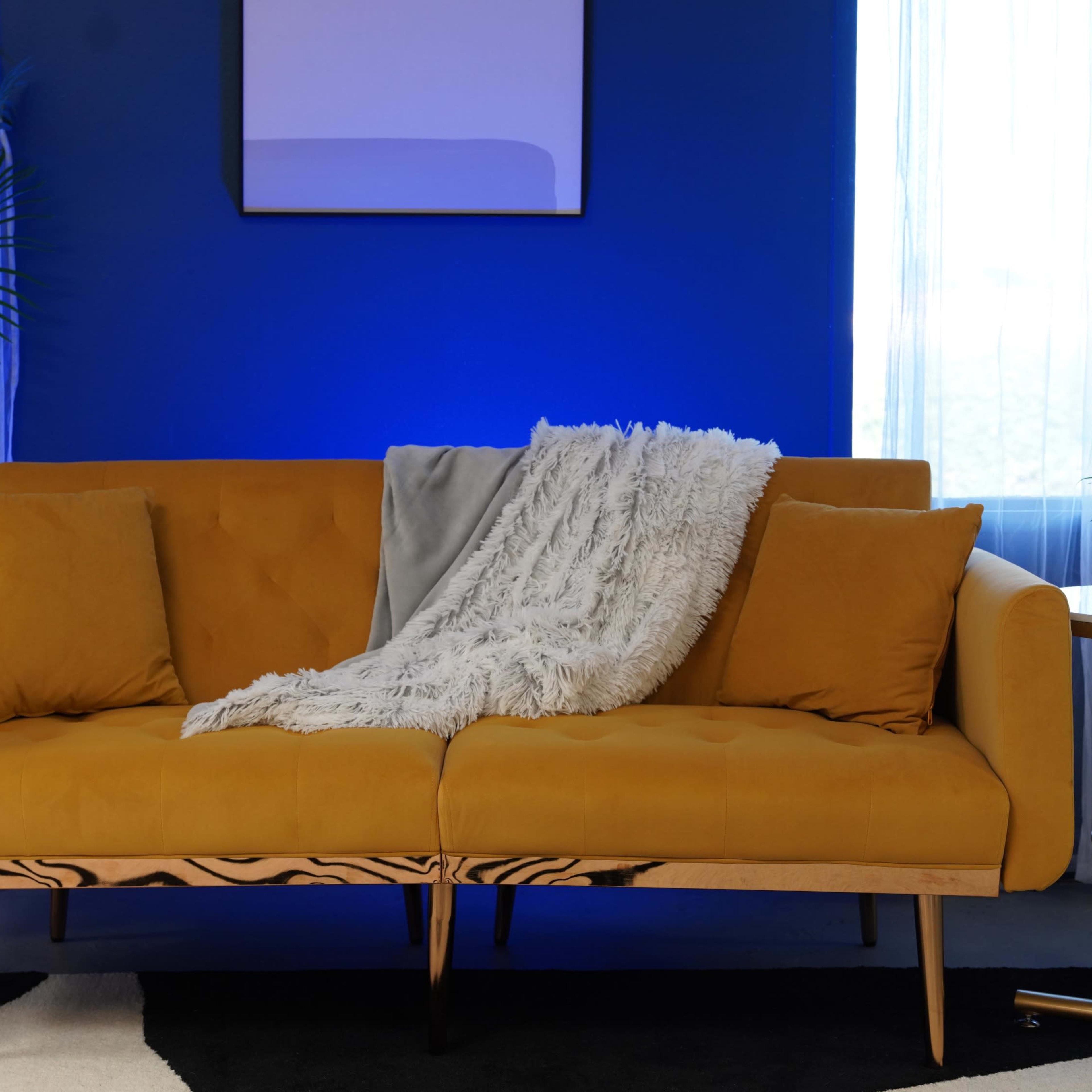 A mustard yellow sofa with a white throw blanket is positioned against a blue wall, accompanied by a plant and a small side table.